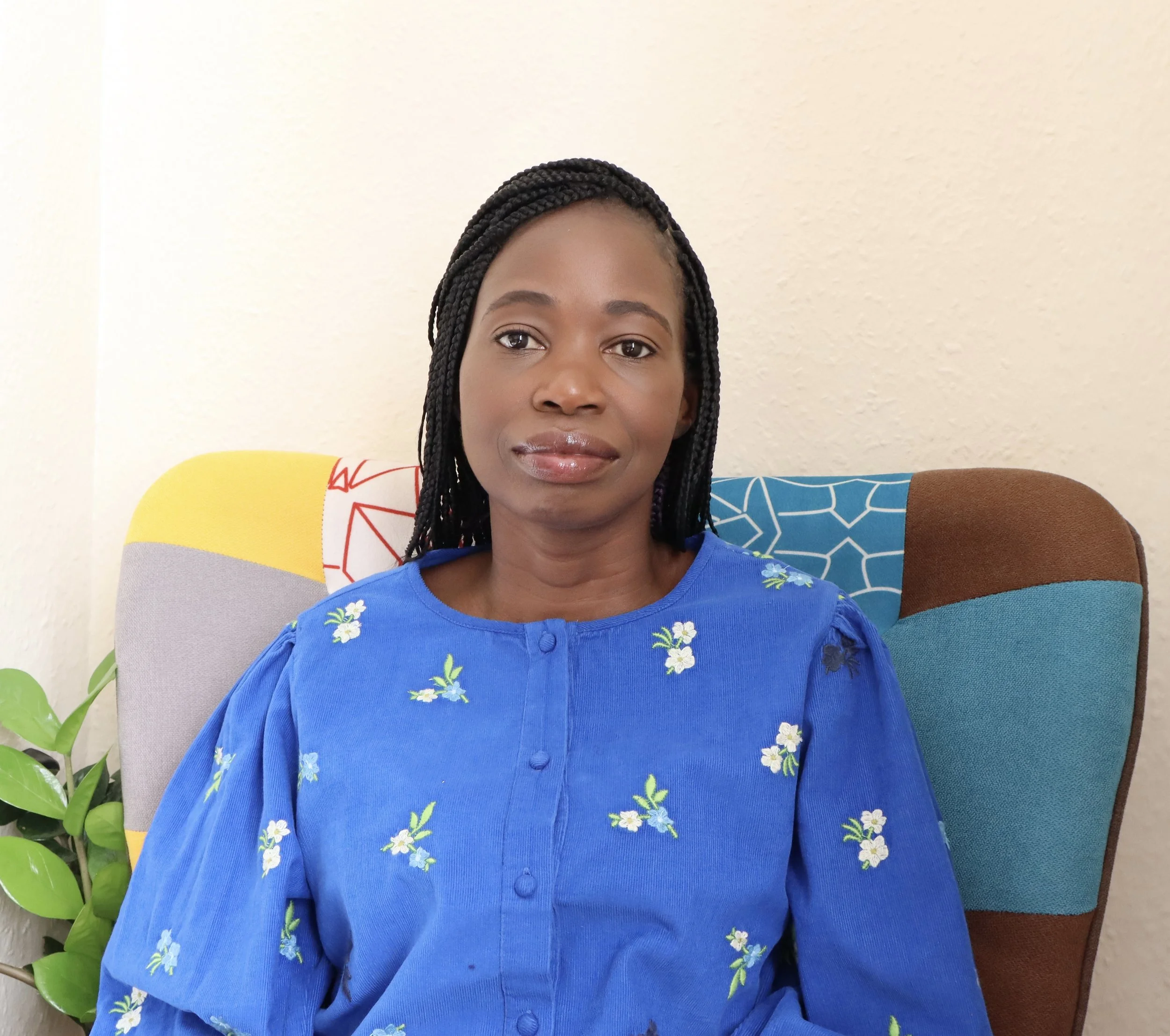 A woman with braided hair sitting on a multicolored couch with a cream wall behind her.