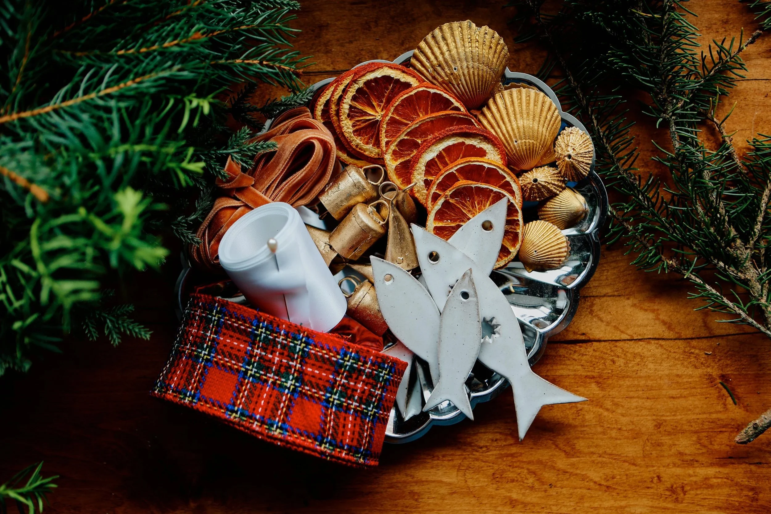Decorative holiday tray with dried orange slices, seashells, small bells, and festive ornaments, surrounded by pine branches on a wooden surface.