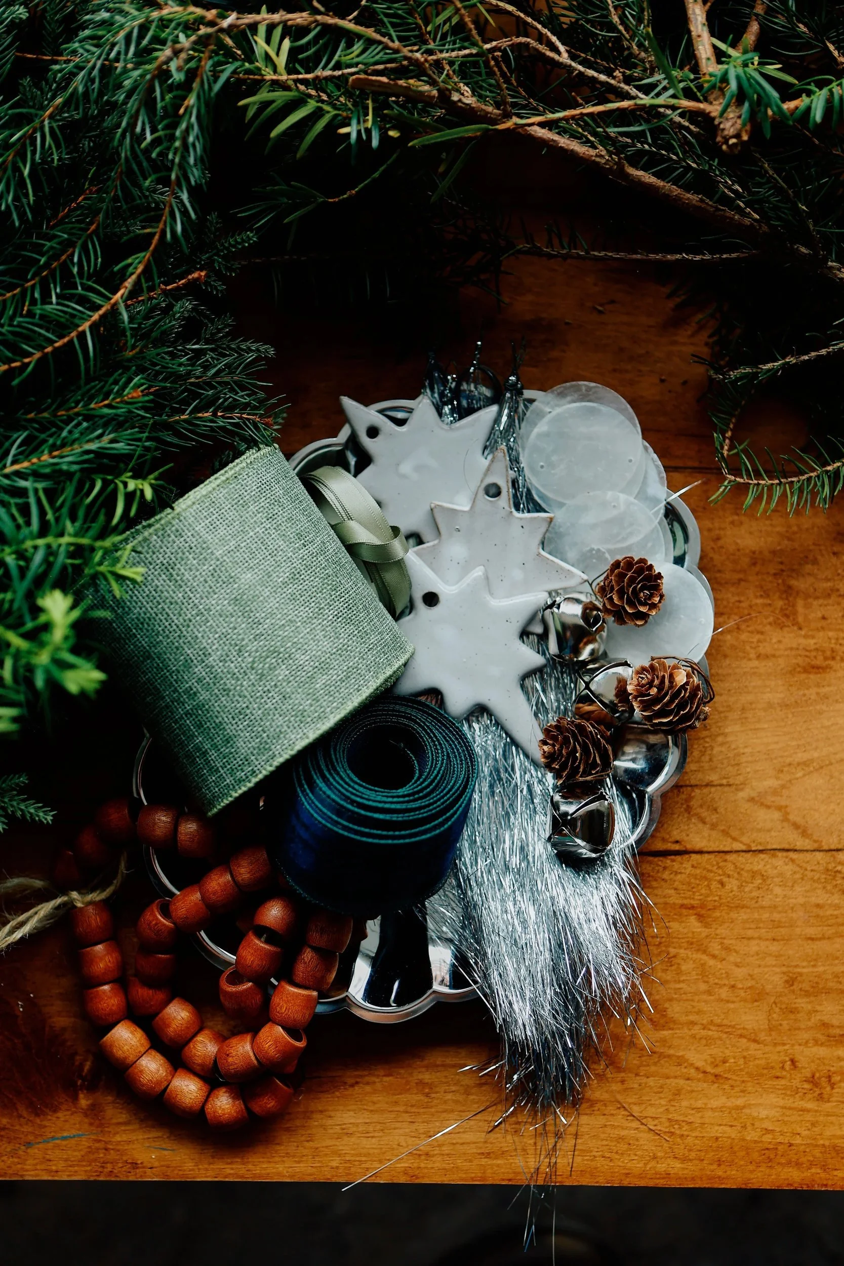Christmas or holiday decorations including pine branches, pinecones, a green fabric ornament, blue ribbon, wooden beads, and a metallic silver star-shaped ornament on a wooden surface.