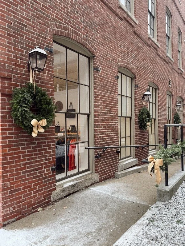 Decorated brick exterior with Christmas wreaths, gold ribbons, and outdoor lanterns, along with a storefront window showing clothing inside.