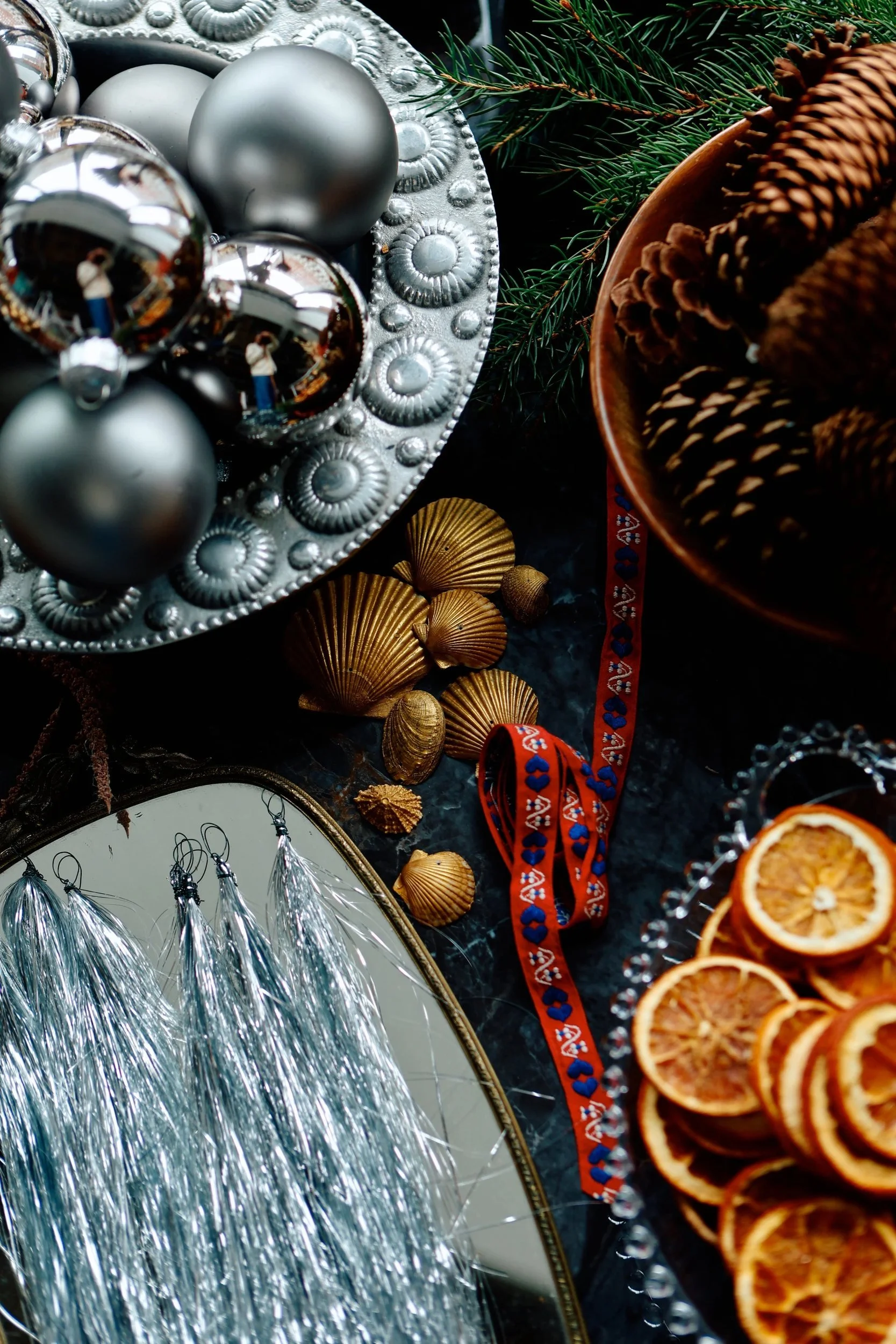 Silver Christmas ornaments, golden shells, dried orange slices, evergreen branches, and decorative ribbons on a dark background.