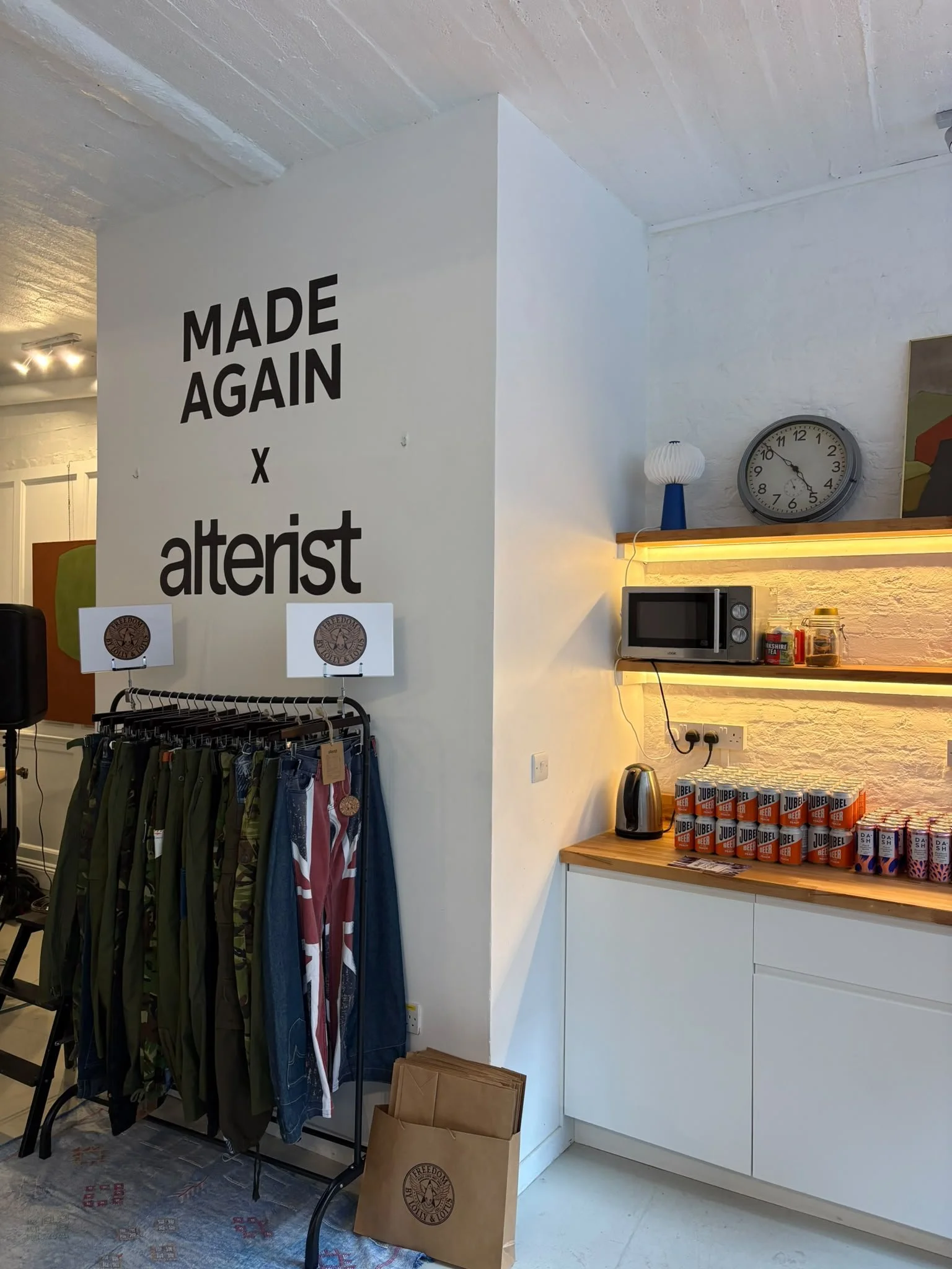 Interior of a store with a white wall featuring a black sign that reads 'Made Again x afterist', clothing on a stand, a microwave oven on a wooden shelf with lighting underneath, a microwave oven, a clock on the wall, a lamp, a kettle, and jars of te