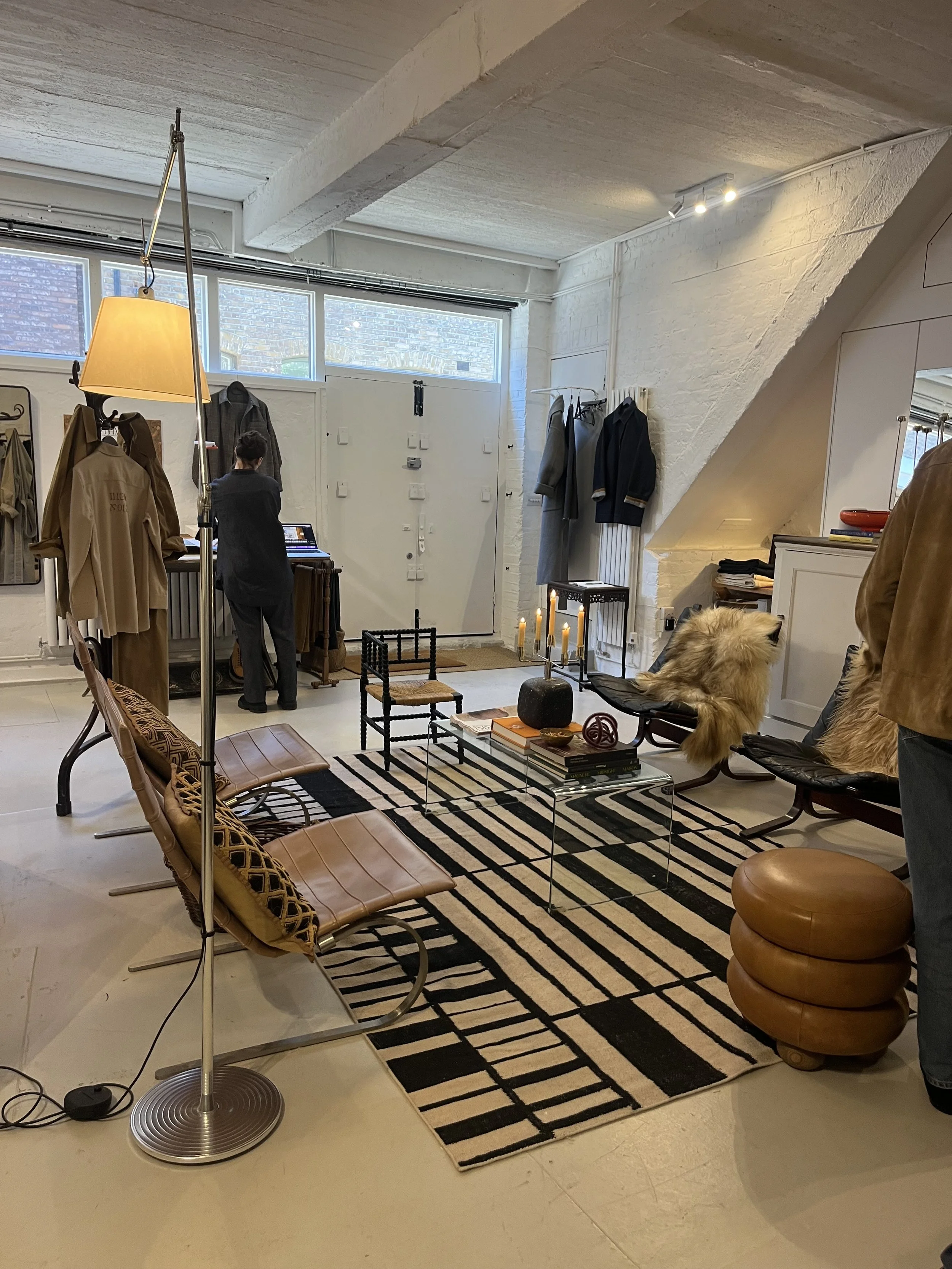 A modern living space with white brick walls, a black and white striped rug, and various seating options including a tan leather chair, a round stacked leather stool, and black chairs. Decor includes a table with books, candles, and artwork, with clo