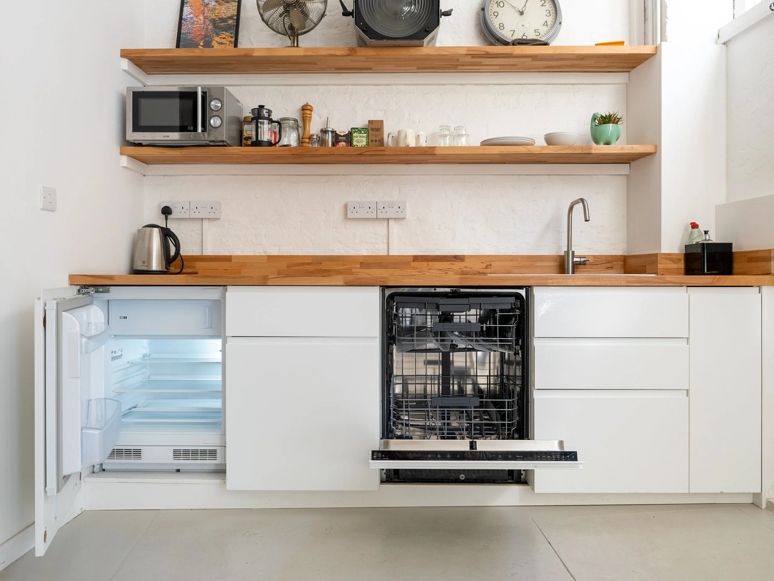 View of a minimalist kitchen with open wooden shelves holding appliances, jars, and a plant, white cabinetry, an open fridge, and a countertop with a sink and black dish rack.