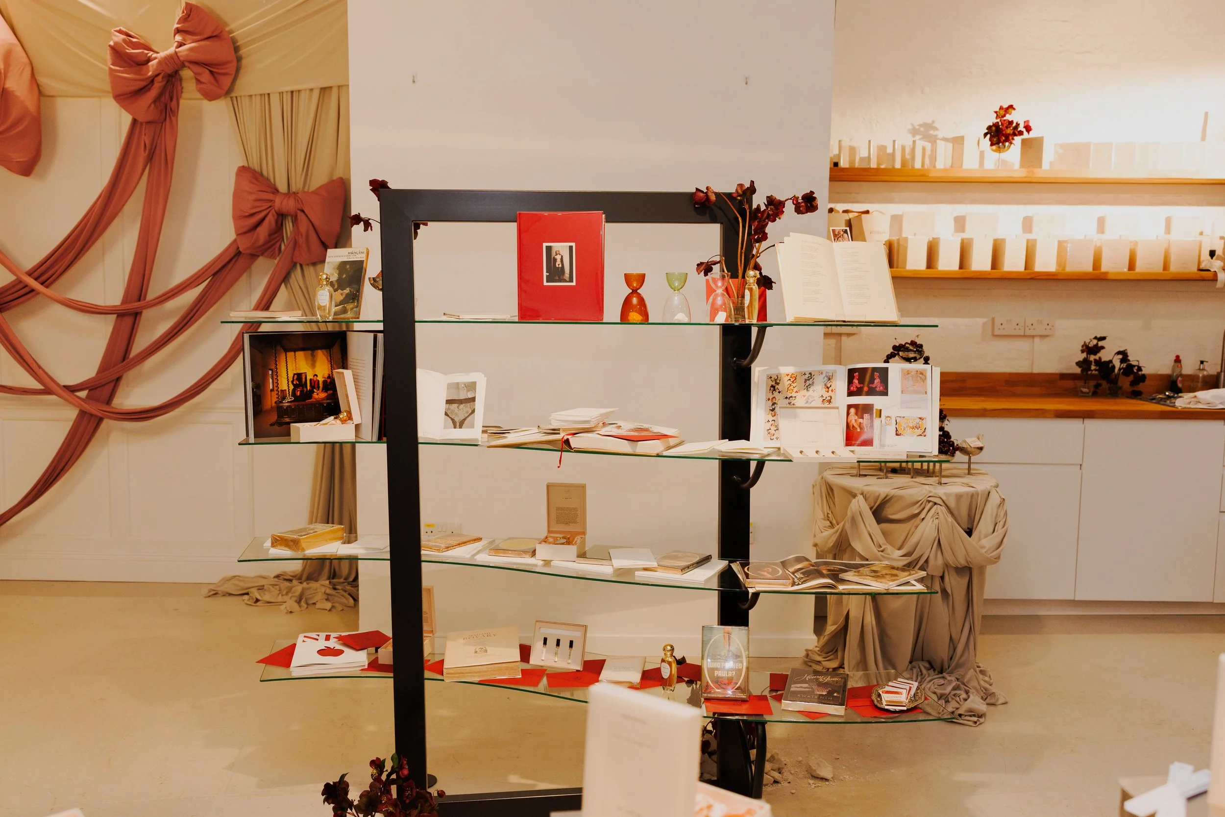 Display stand with art books, magazines, decorative glassware, and floral arrangements in a gallery setting.