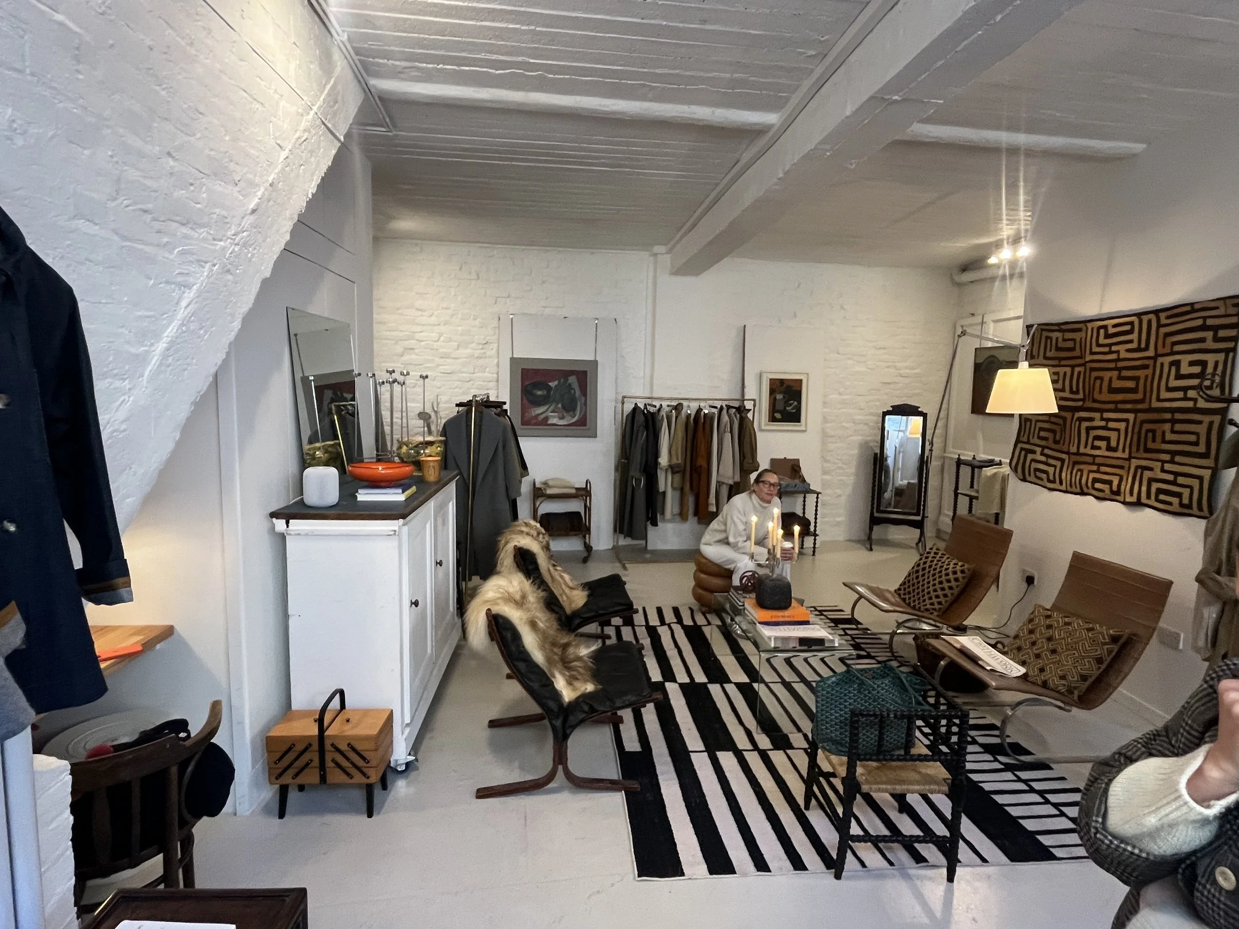 Interior of a modern, stylish boutique or showroom with white brick walls, a black and white striped rug, and various clothing items on racks and chairs. There is a woman sitting on a chair holding a candle, a large sofa with decorative pillows, and 