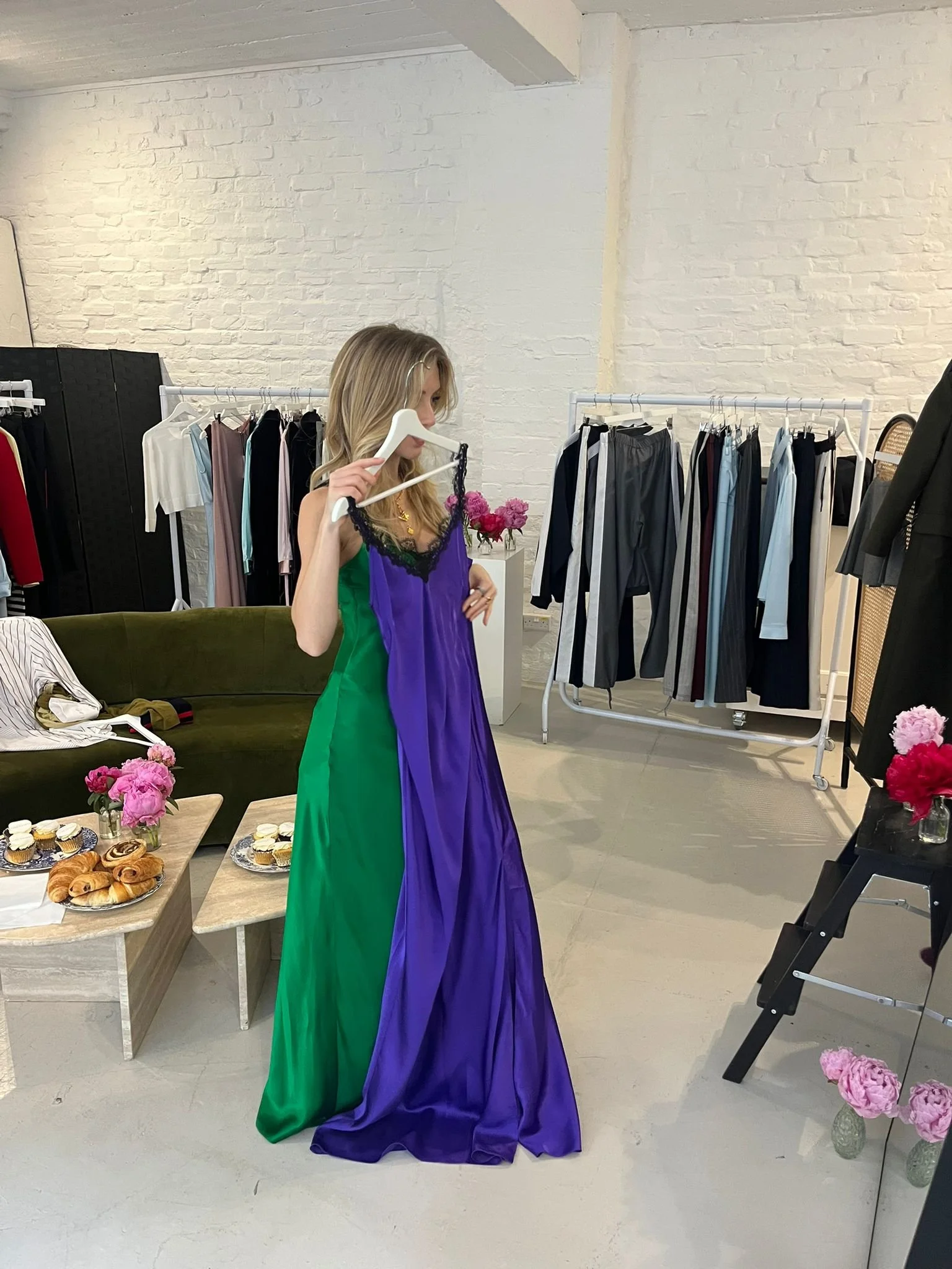 A woman in a green and purple dress holds a purple dress on a hanger in a boutique or dressing room with white brick walls. There are clothing racks with pants and tops, a table with pink flowers, pastries, and a black clothing rack with additional g