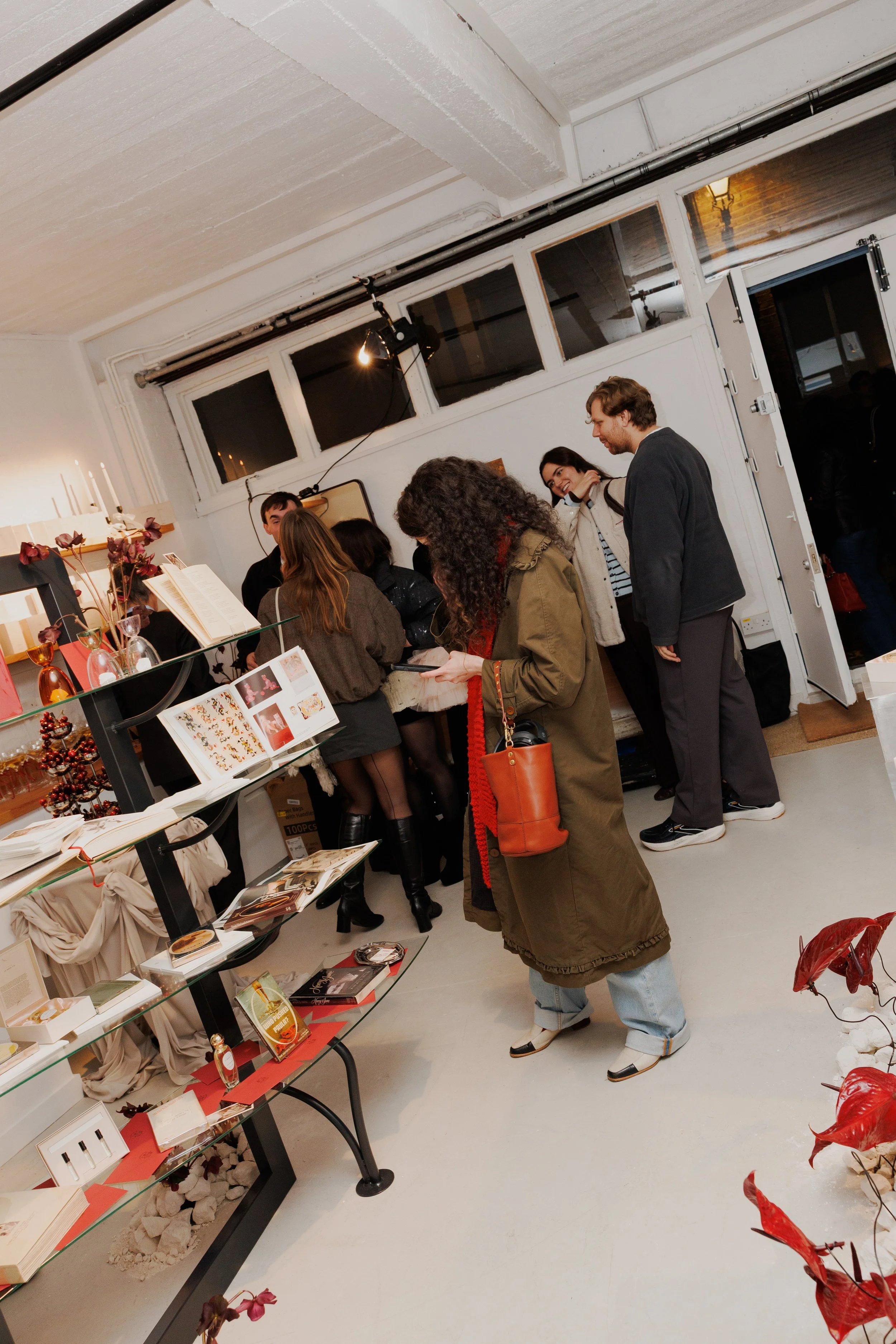 People shopping at an indoor store, browsing items on display shelves with books, decor, and ornamental objects, near a wall with windows.