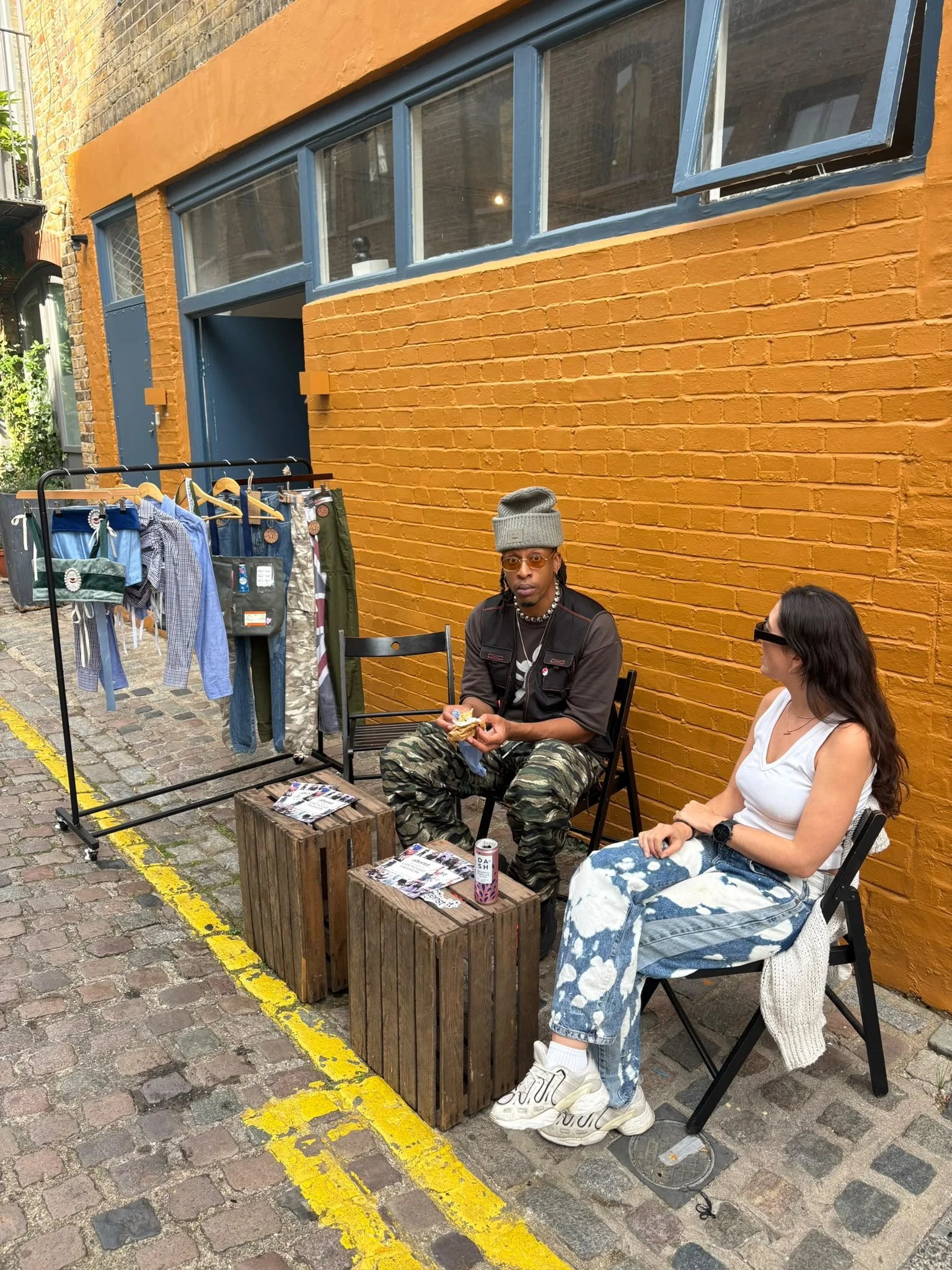 chairs outside against an orange brick wall, with a clothing rack displaying men's clothes nearby. The woman on the left wears a gray beanie, glasses, and camouflage pants, and is eating. The woman on the right wears a white tank top, tie-dye jeans, 