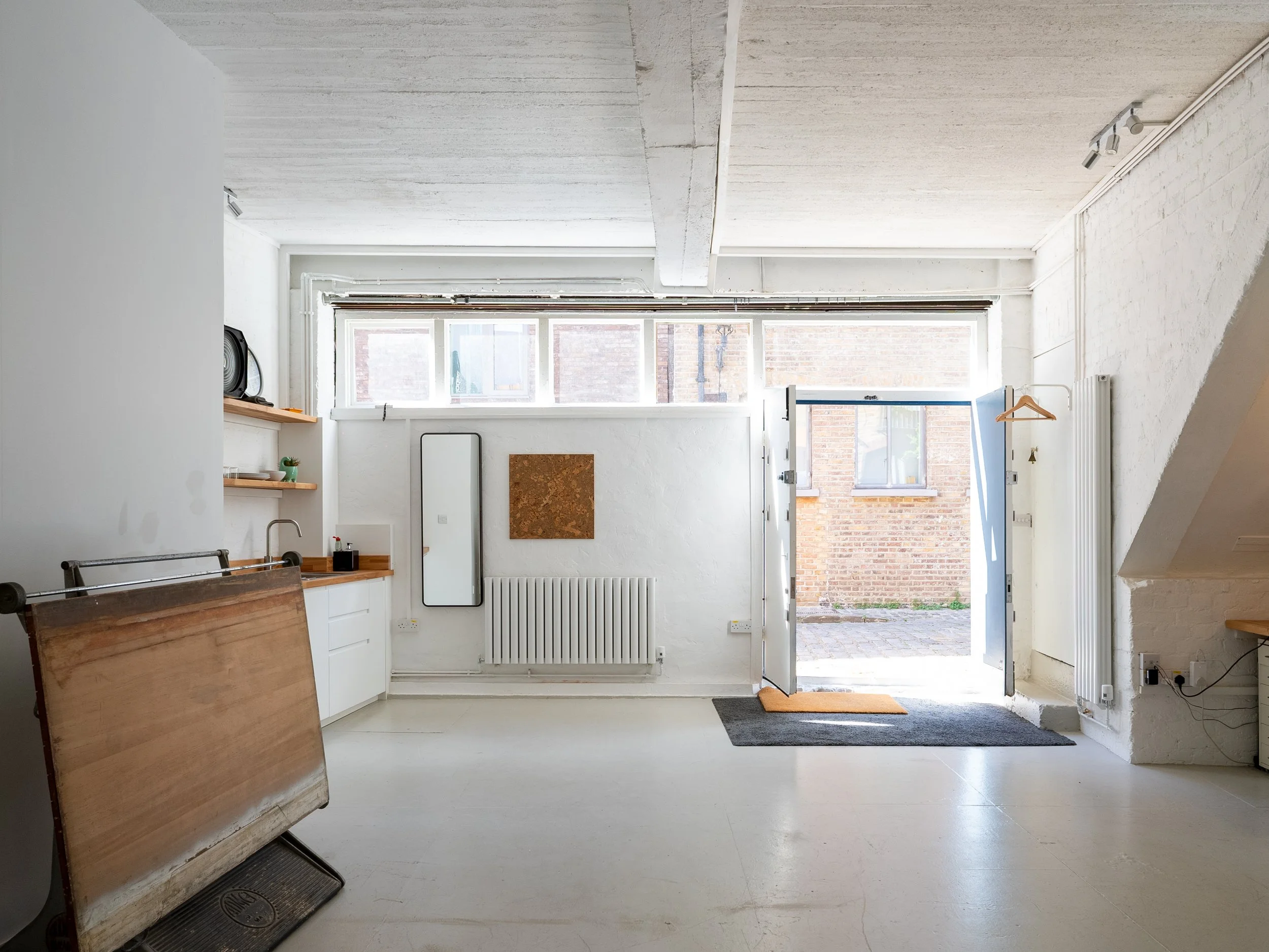 Interior view of a minimalist room with white walls and ceiling, a large window letting in natural light, a radiator beneath the window, a chair with wooden slats leaning against the wall, shelves with decorative items, a mirror, and an open door leading outside with visible brick buildings and pavement.