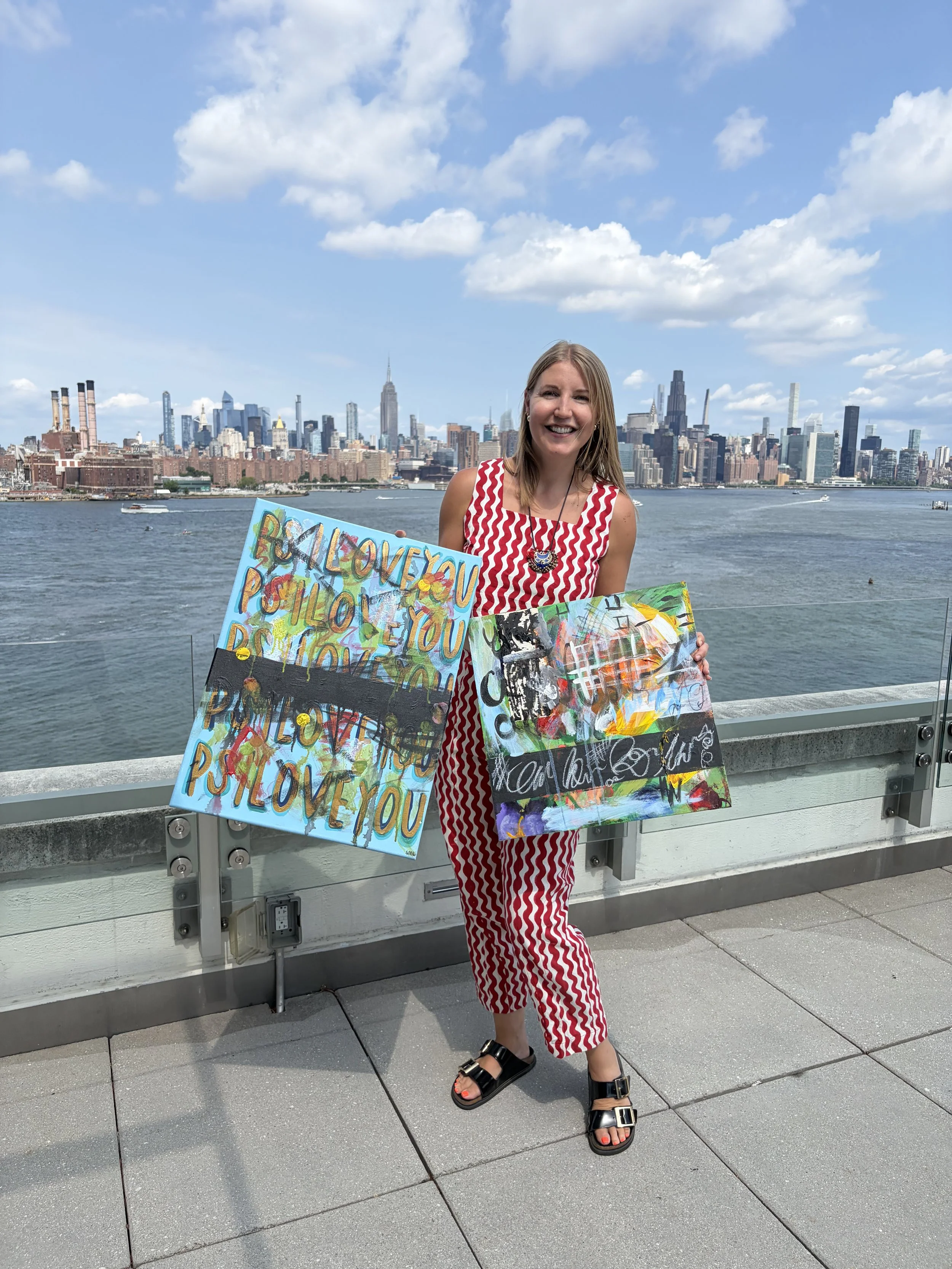 Summer Lindman holding two paintings in front of the New York City skyline.