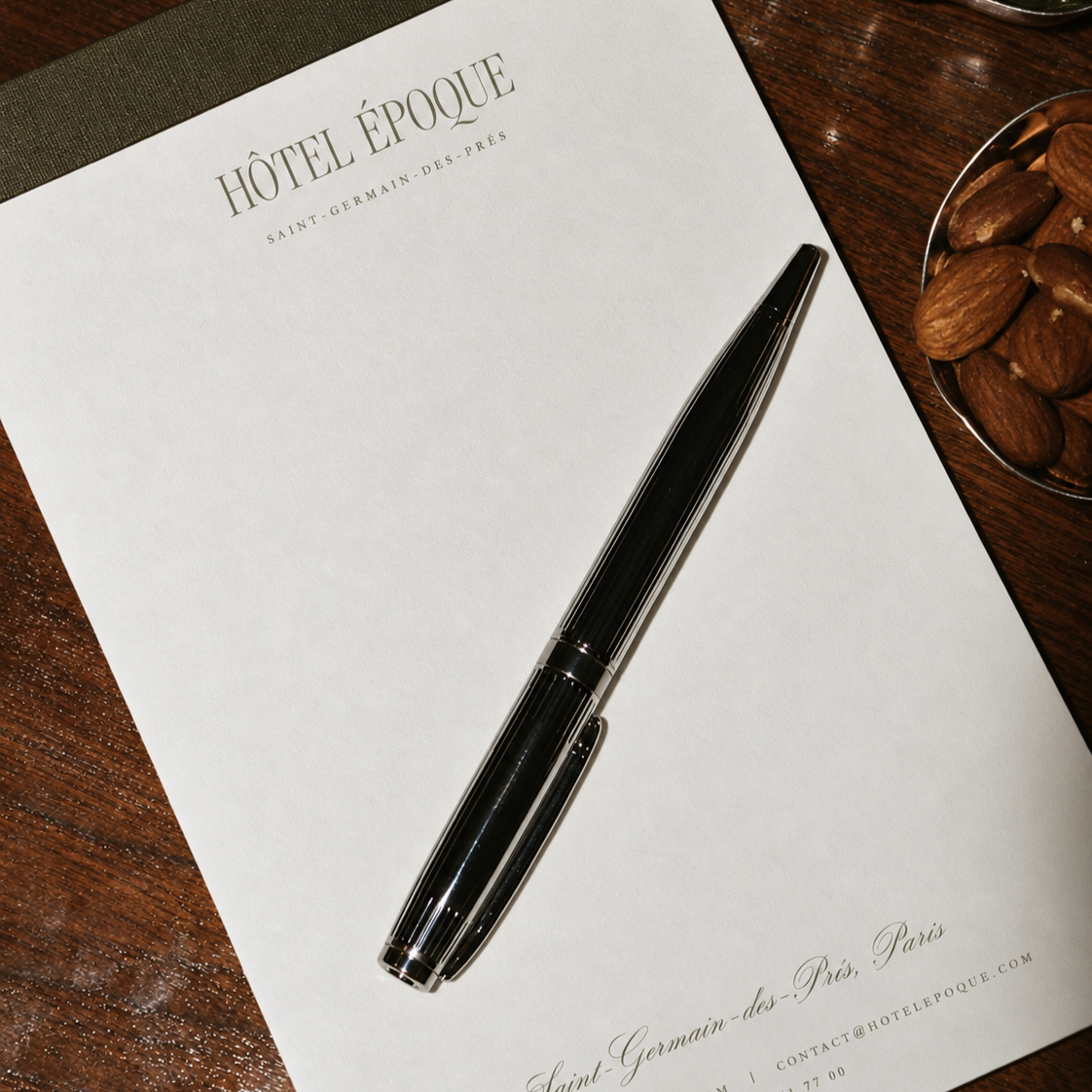 A black pen resting on a white notepad with 'Hôtel Époque' printed at the top and contact information at the bottom, placed on a wooden surface beside a bowl of almonds.