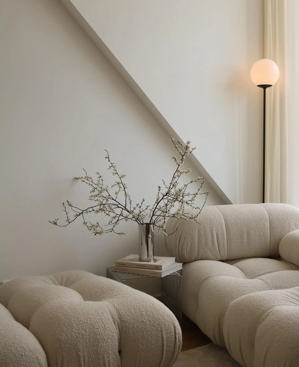 Minimalist living room corner with a beige sofa, white wall, a side table with a stack of books, a silver vase with branches, and a tall black floor lamp with a round white lampshade near cream curtains.