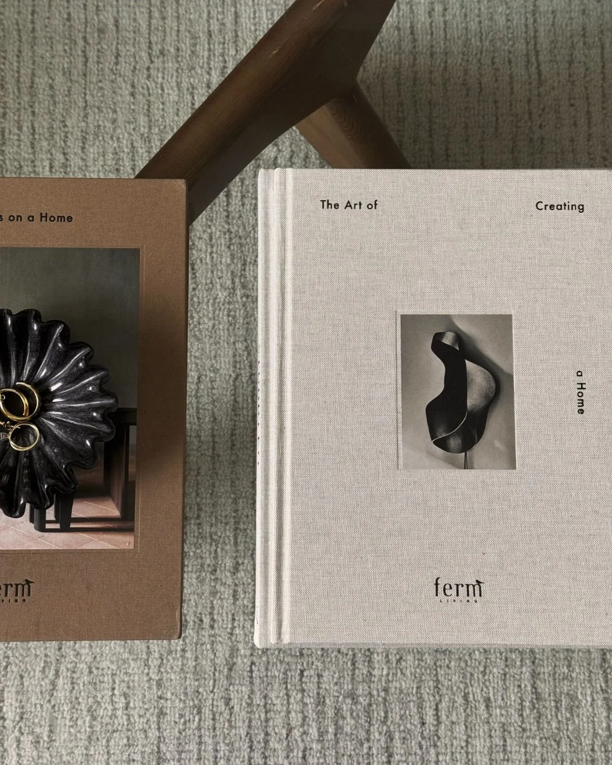 Two books on art and home decor placed on a textured beige carpet, with a small black decorative object in a black frame partially visible behind the brown book.