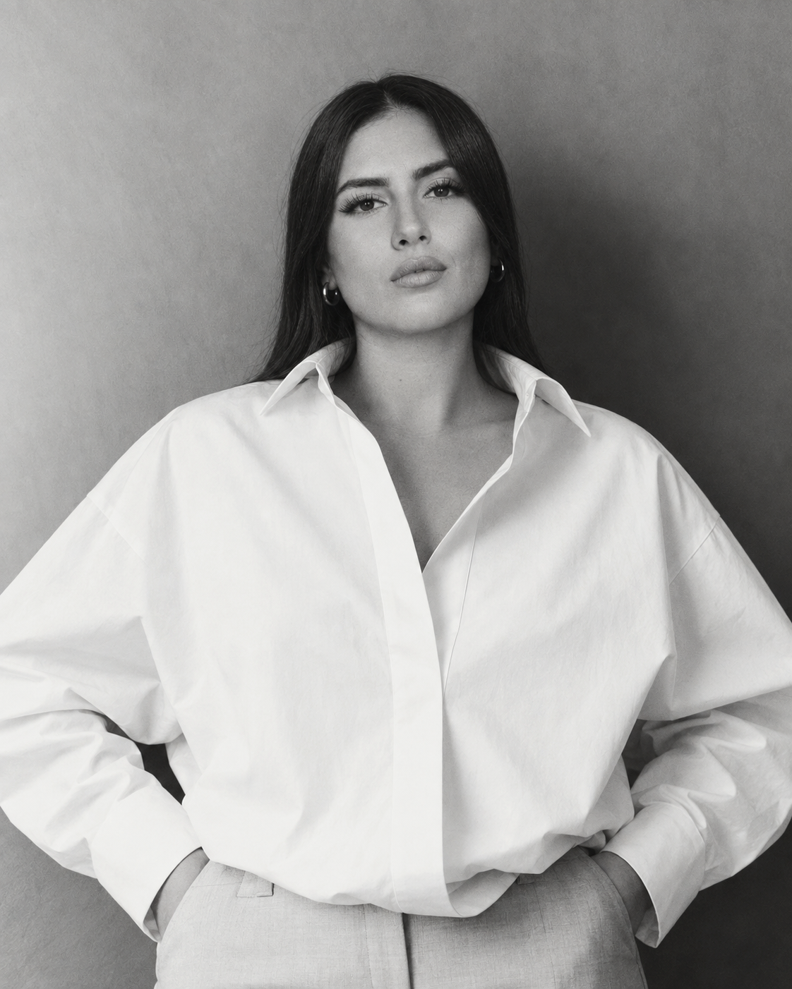 A woman with long dark hair and hoop earrings, wearing a white oversized dress shirt, standing against a plain wall.