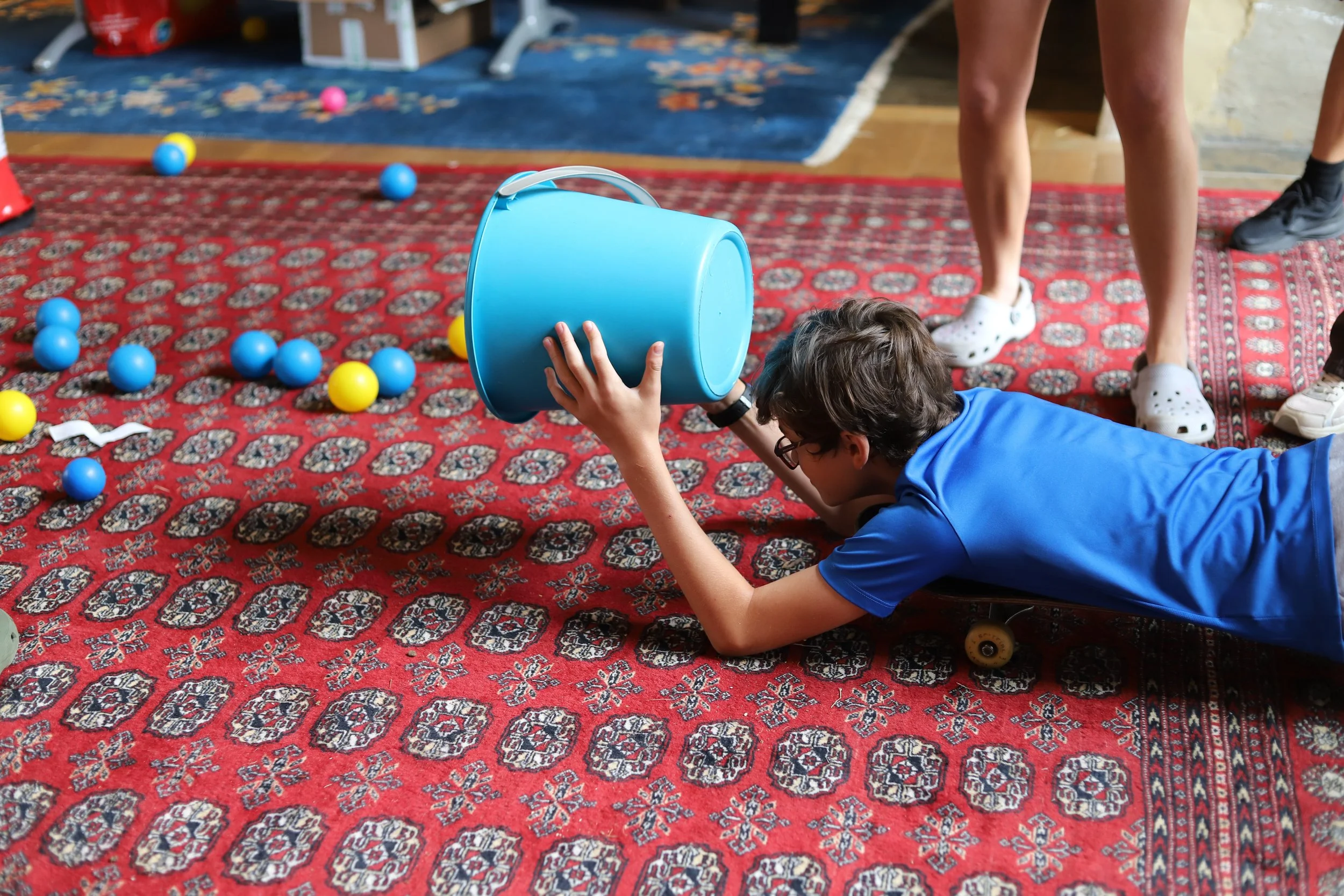 Teenage boy collects pit balls with a bucket while lying on a skateboard.