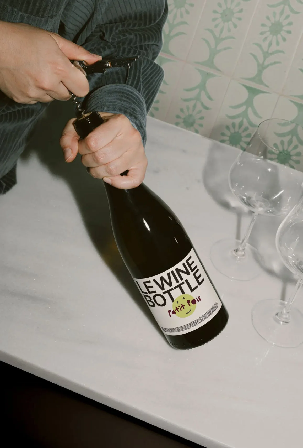 Henkilö avaa pulloa, jonka etiketti lukee 'Le Wine Bottle' ja 'Petit Rosi', vieressä on tyhjiä viinilaseja pöydällä.