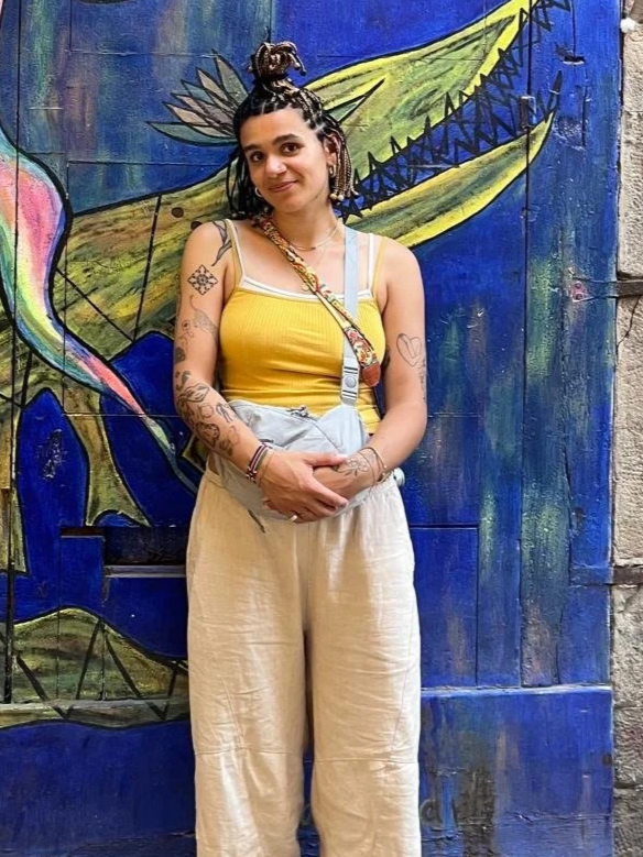 A woman with braided hair styled in an updo, wearing a yellow tank top and beige wide-leg pants, standing in front of a colorful mural with abstract designs and a dragonfly.