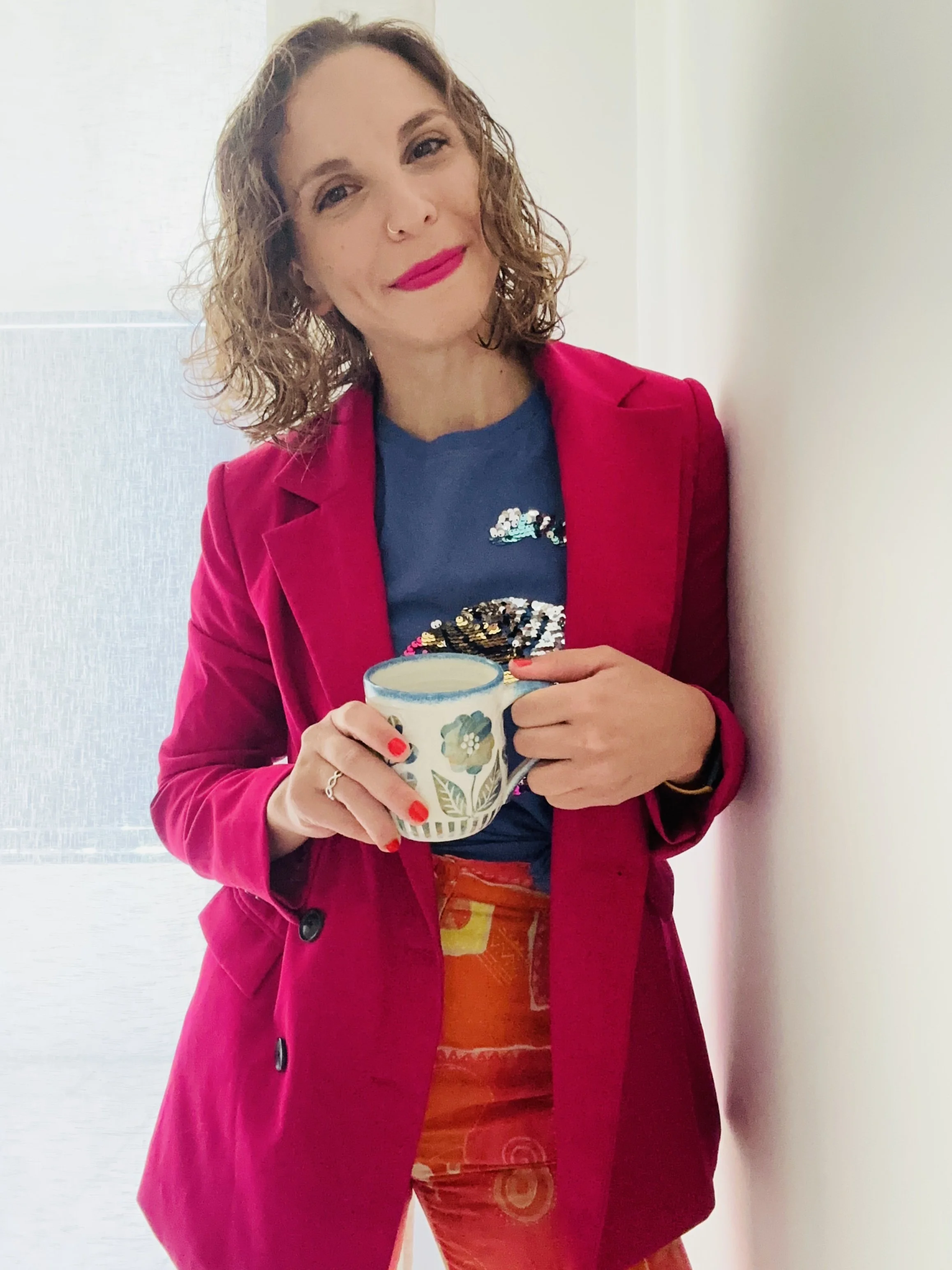 A woman with curly hair, pink lipstick, wearing a red blazer, and a blue top with sequins, is smiling and holding a colorful mug with floral and animal patterns. She is standing near a plain wall and a window.