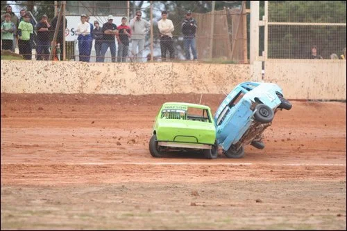 Two small cars crash on a dirt race track, with spectators watching from behind a fence.