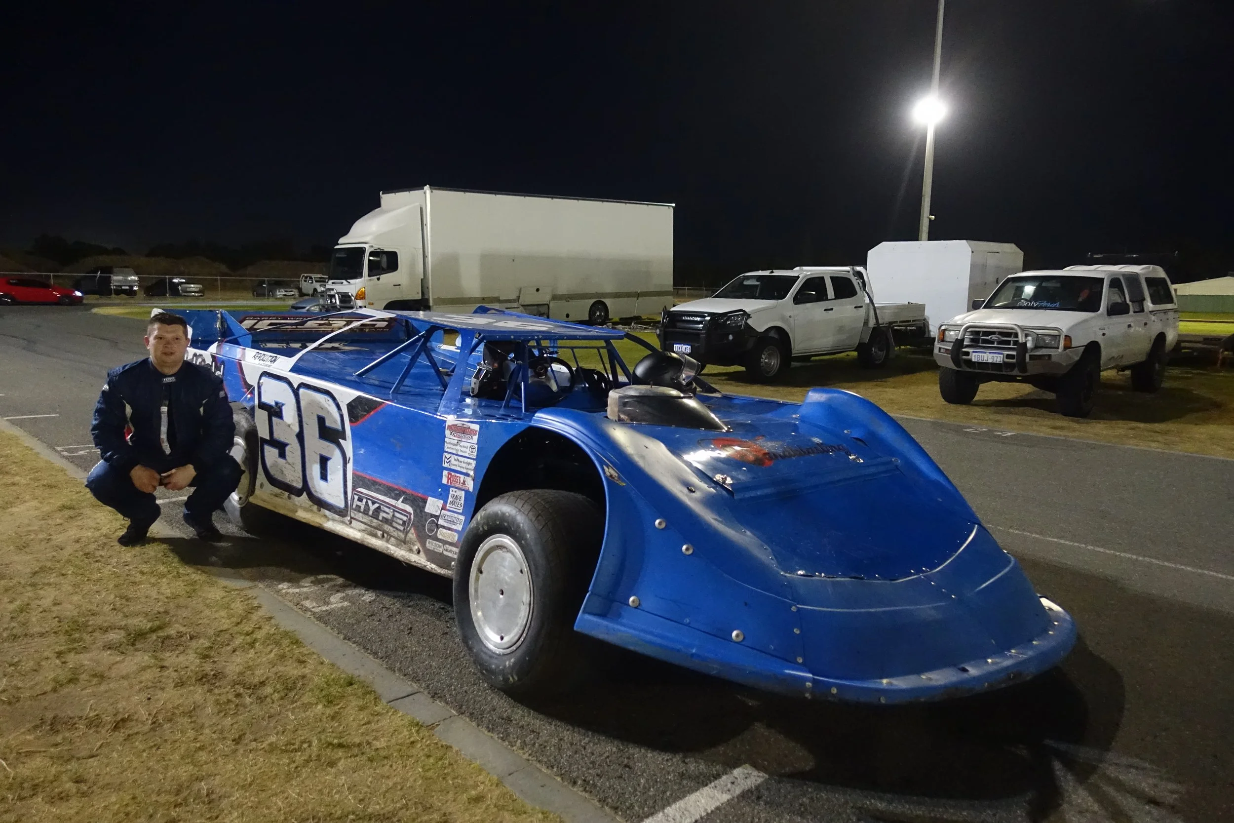 A man in racing suit kneeling beside a blue race car with the number 36, parked in a dimly lit parking lot at night. There are several trucks and vehicles in the background under a tall lamp post.