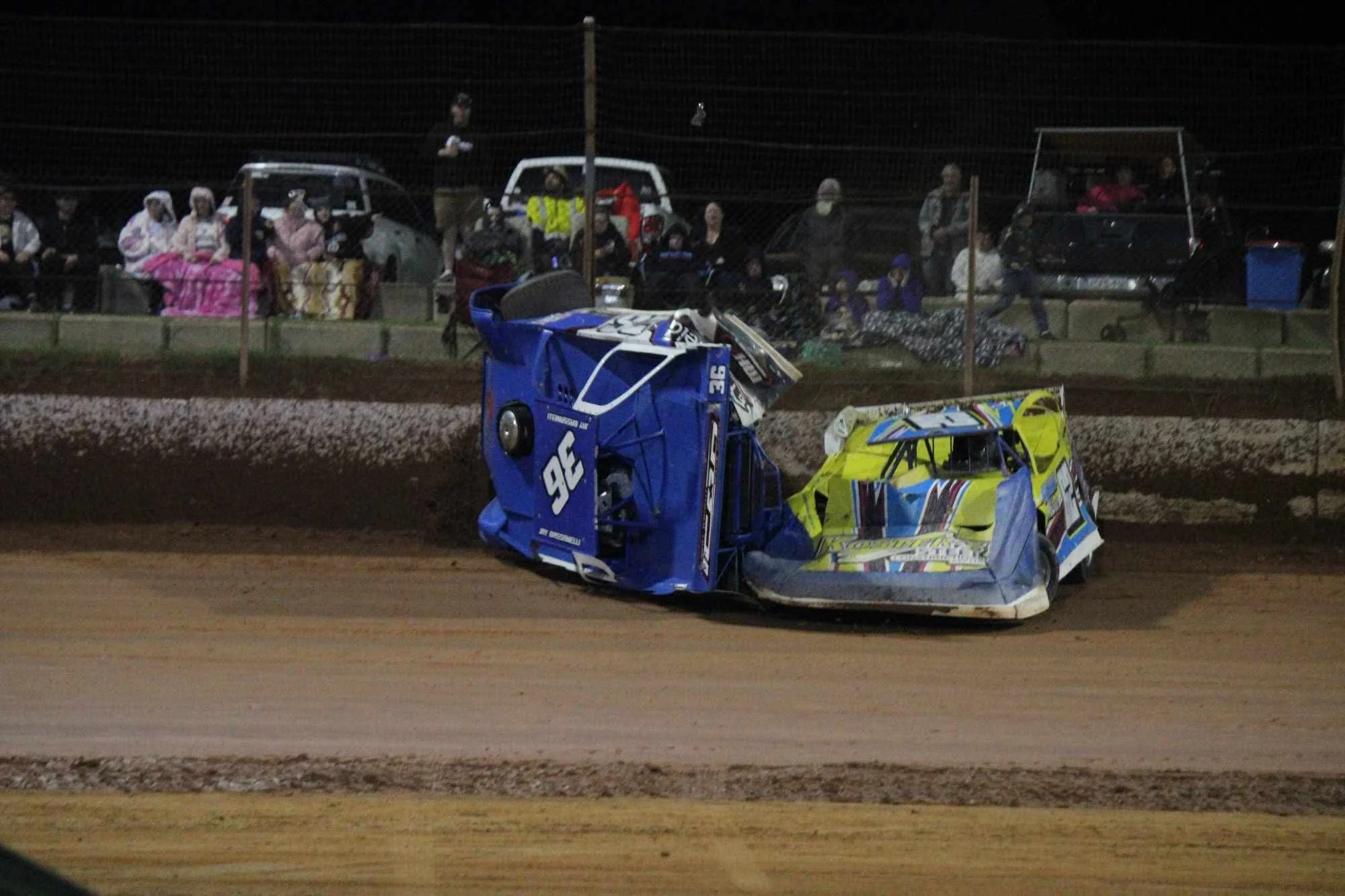 Nighttime dirt track race with wrecked race cars in foreground, crowd of spectators behind a fence, some sitting on blankets, some standing, some in vehicles, watching the race.