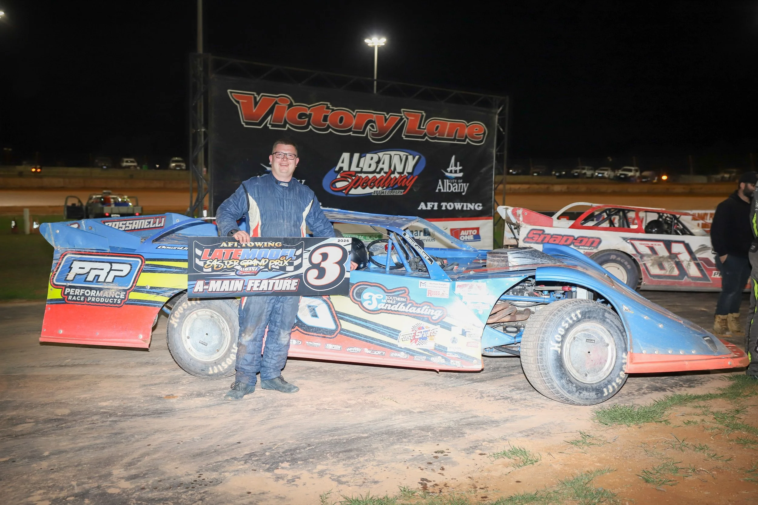 A young man in a racing suit is standing next to a blue race car with various sponsor decals at a dirt racetrack during nighttime. He is smiling and holding a sign that promotes a late model racing event at Albany Speedway.