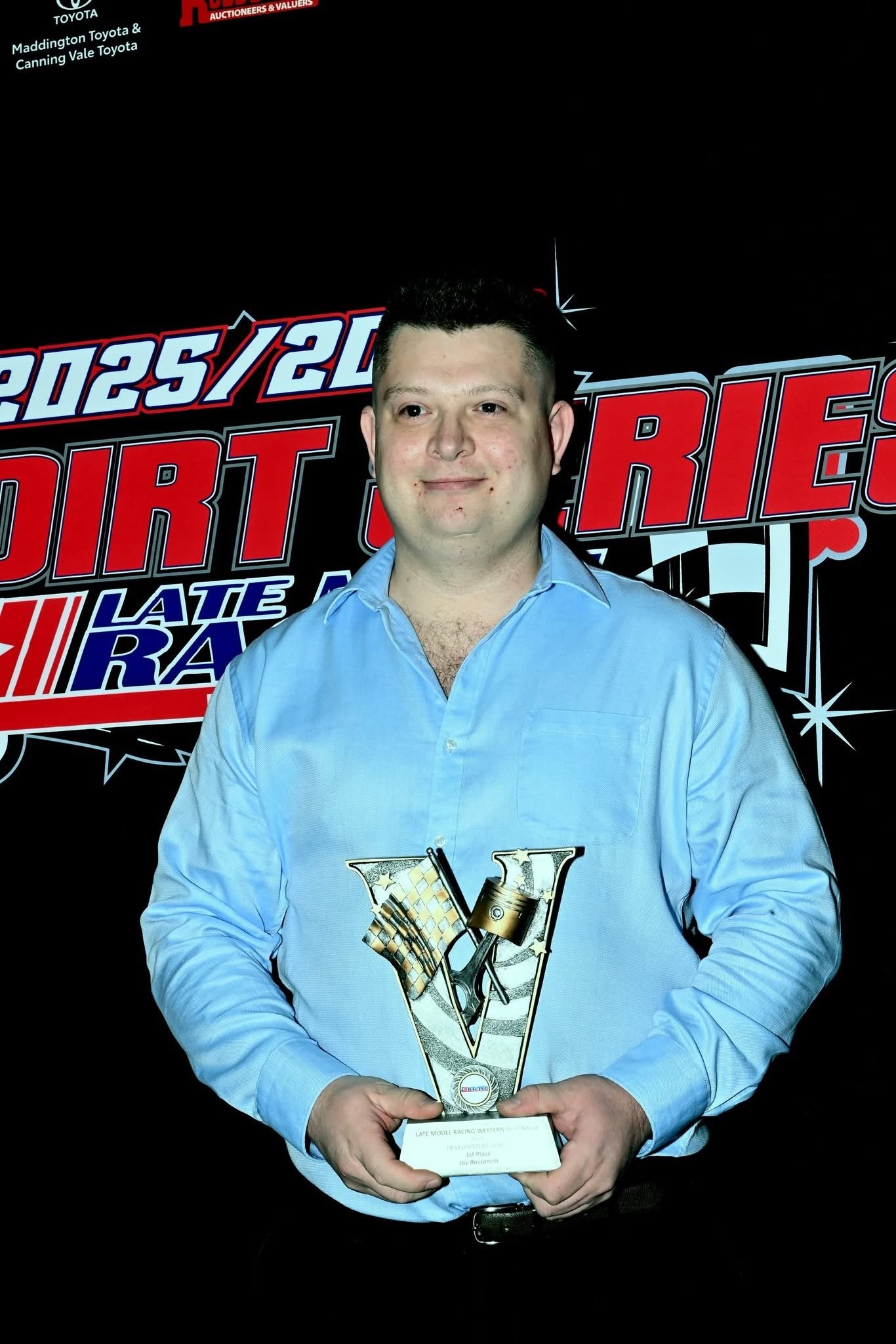 A man in a light blue shirt holding a trophy in front of a black background with racing-related logos and text.