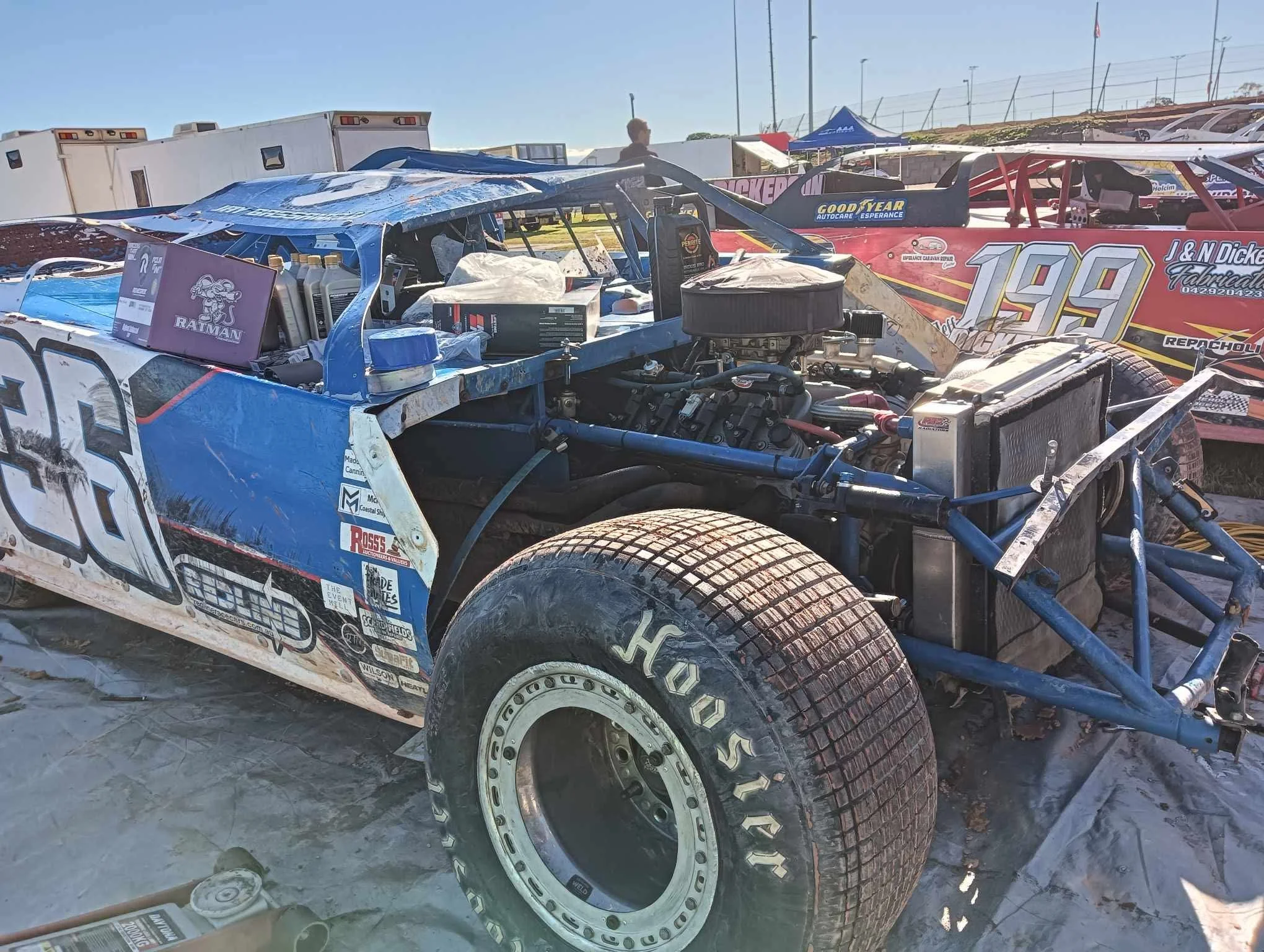 A damaged race car with visible engine components and racing stickers on the side, parked outdoors during daytime with other race cars and trailers in the background.