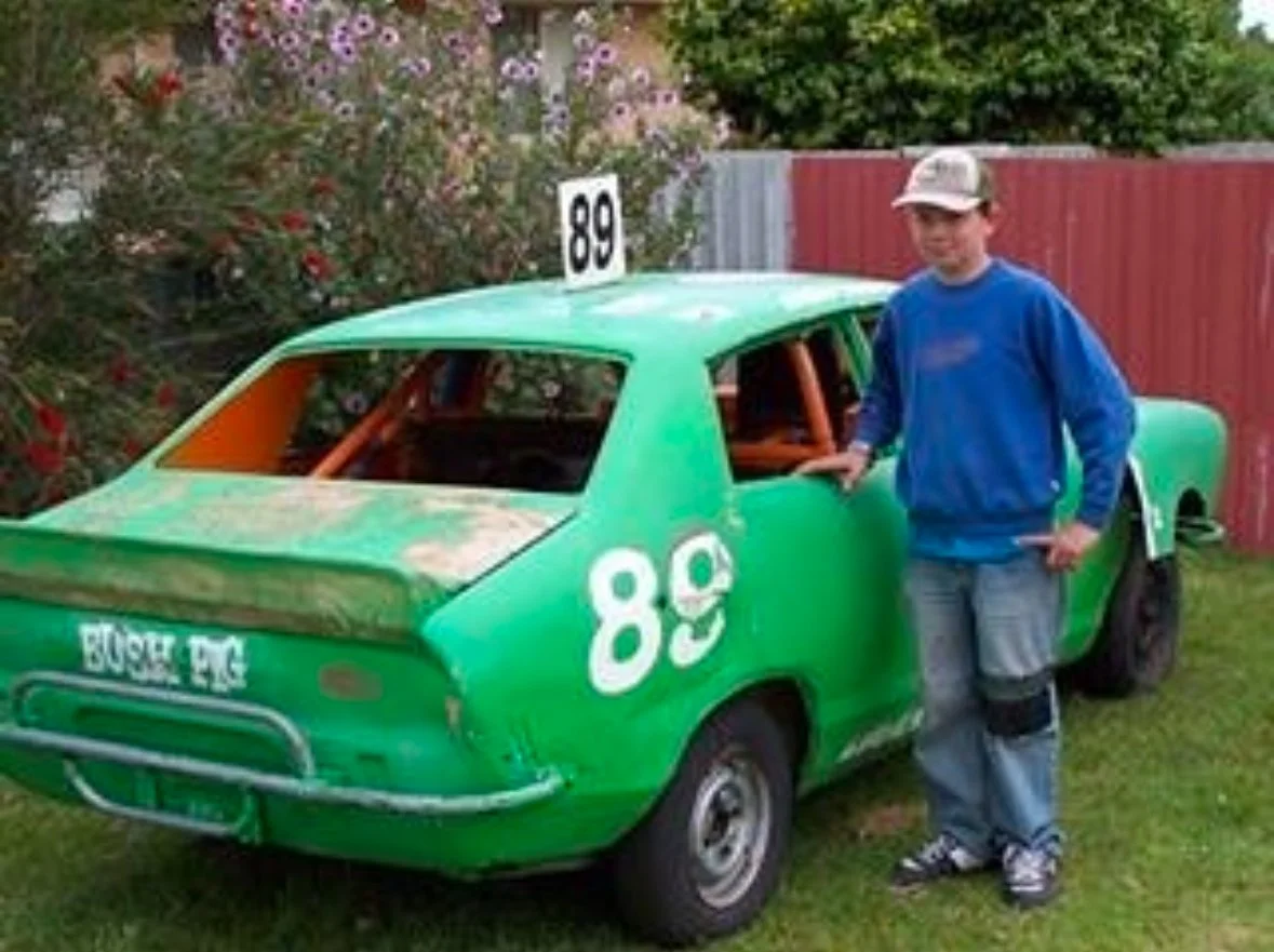 A boy in a blue jacket, gray pants, and a baseball cap standing next to a small green race car with the number 89 and a sign on top, parked on grass in a backyard, with bushes and a red fence in the background.