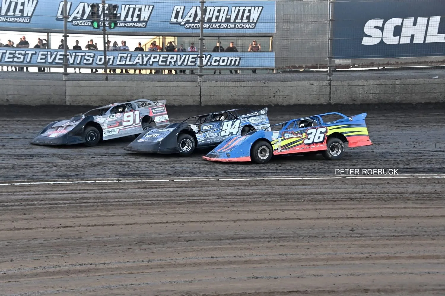 Three race cars on a dirt track racing side by side, with a crowd of spectators and advertising banners in the background.