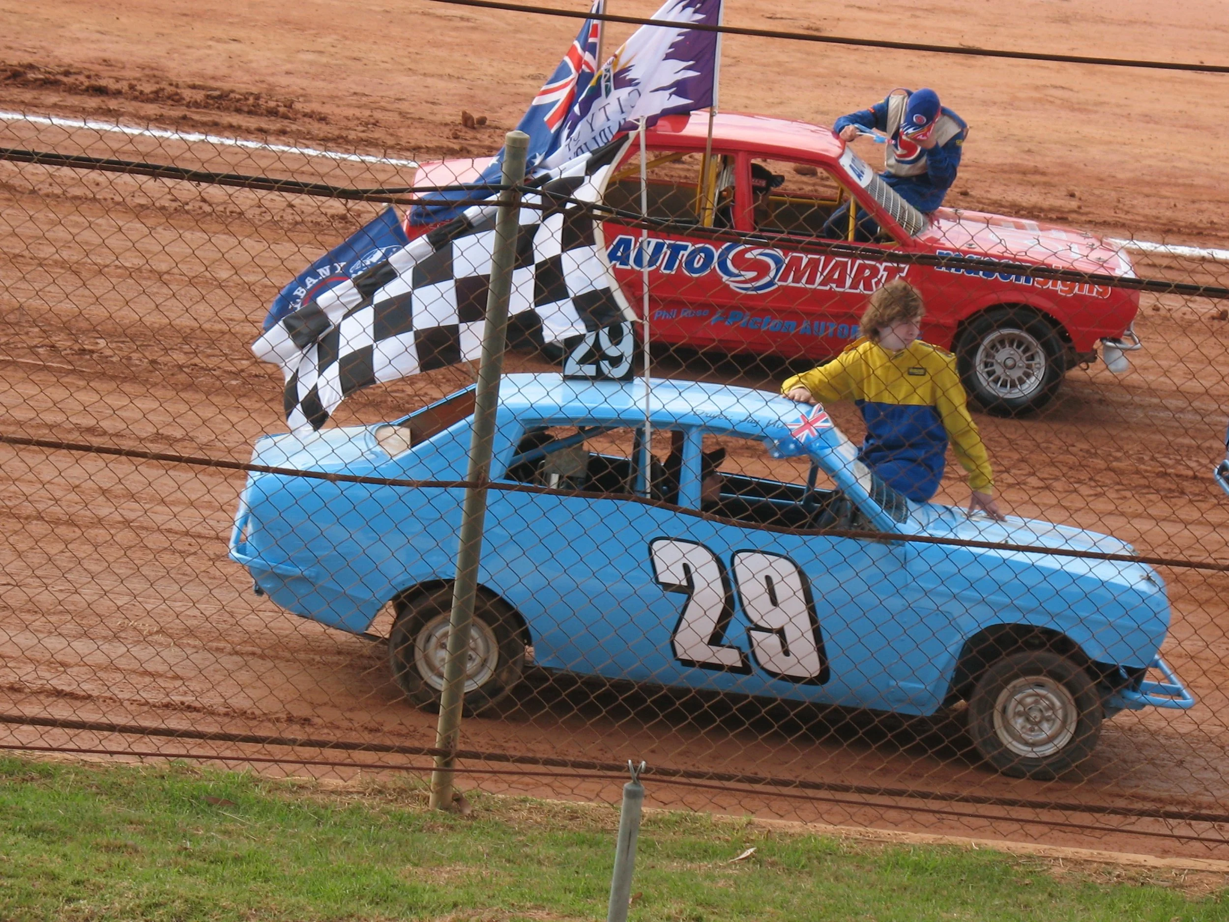Race car with number 29 and event logos, a person in a racing suit working on the car, a person in a yellow jacket standing nearby, on a dirt race track with a wire fence in front.