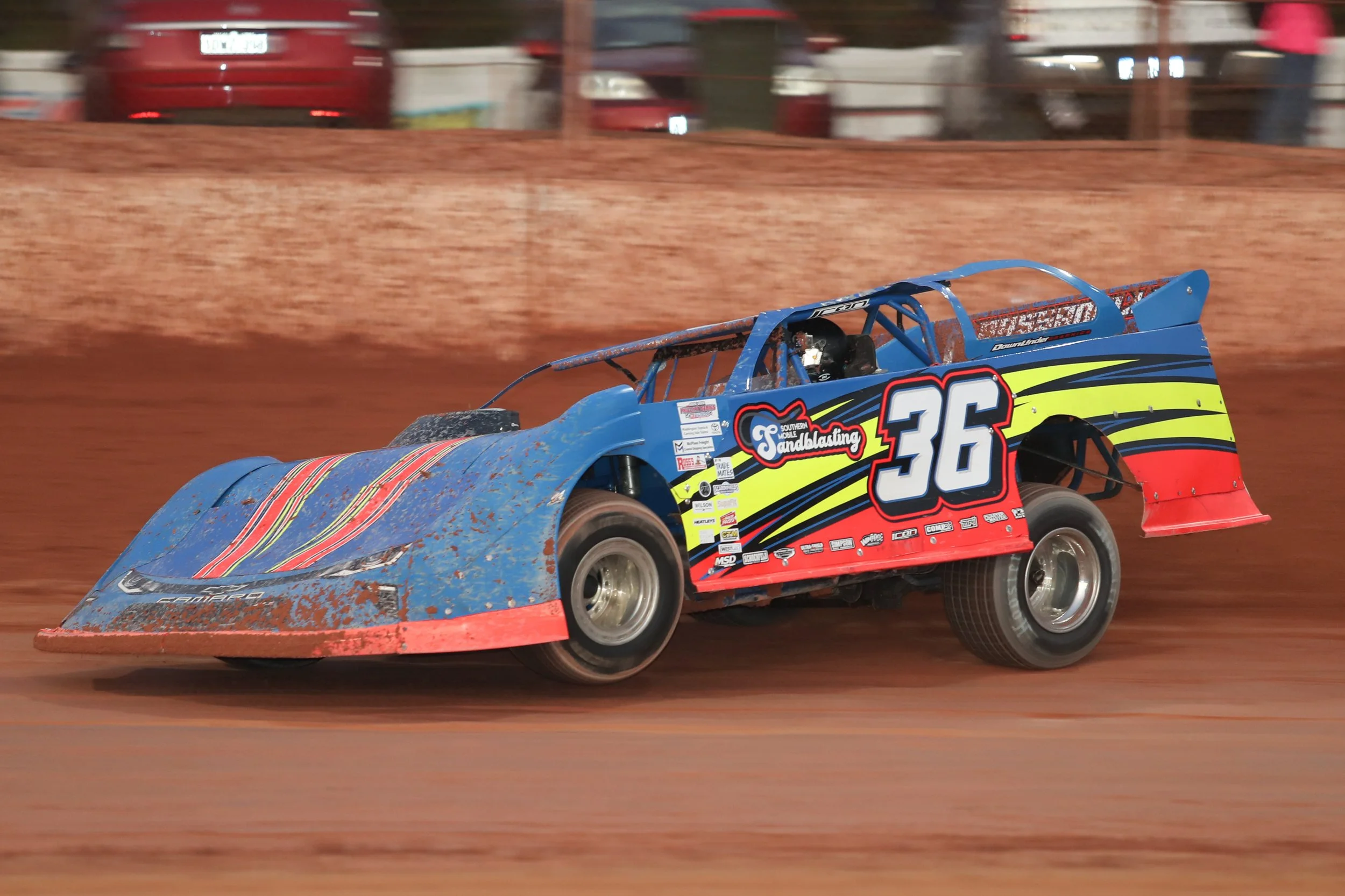 A blue race car with the number 36 driving on a dirt track, kicking up dirt behind it, with a red wall and blurred cars and spectators in the background.
