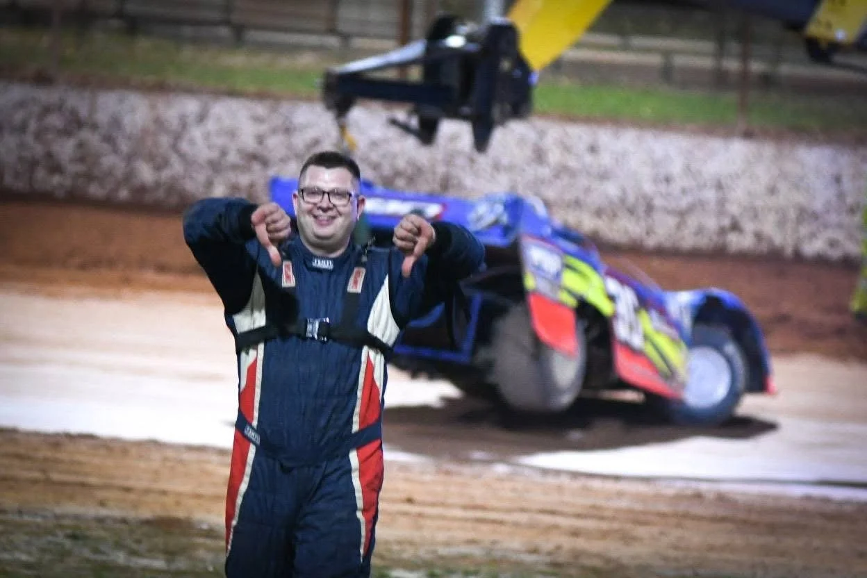 Smiling race car driver in a racing suit giving thumbs down on a dirt track, with a pulled-over race car in the background.