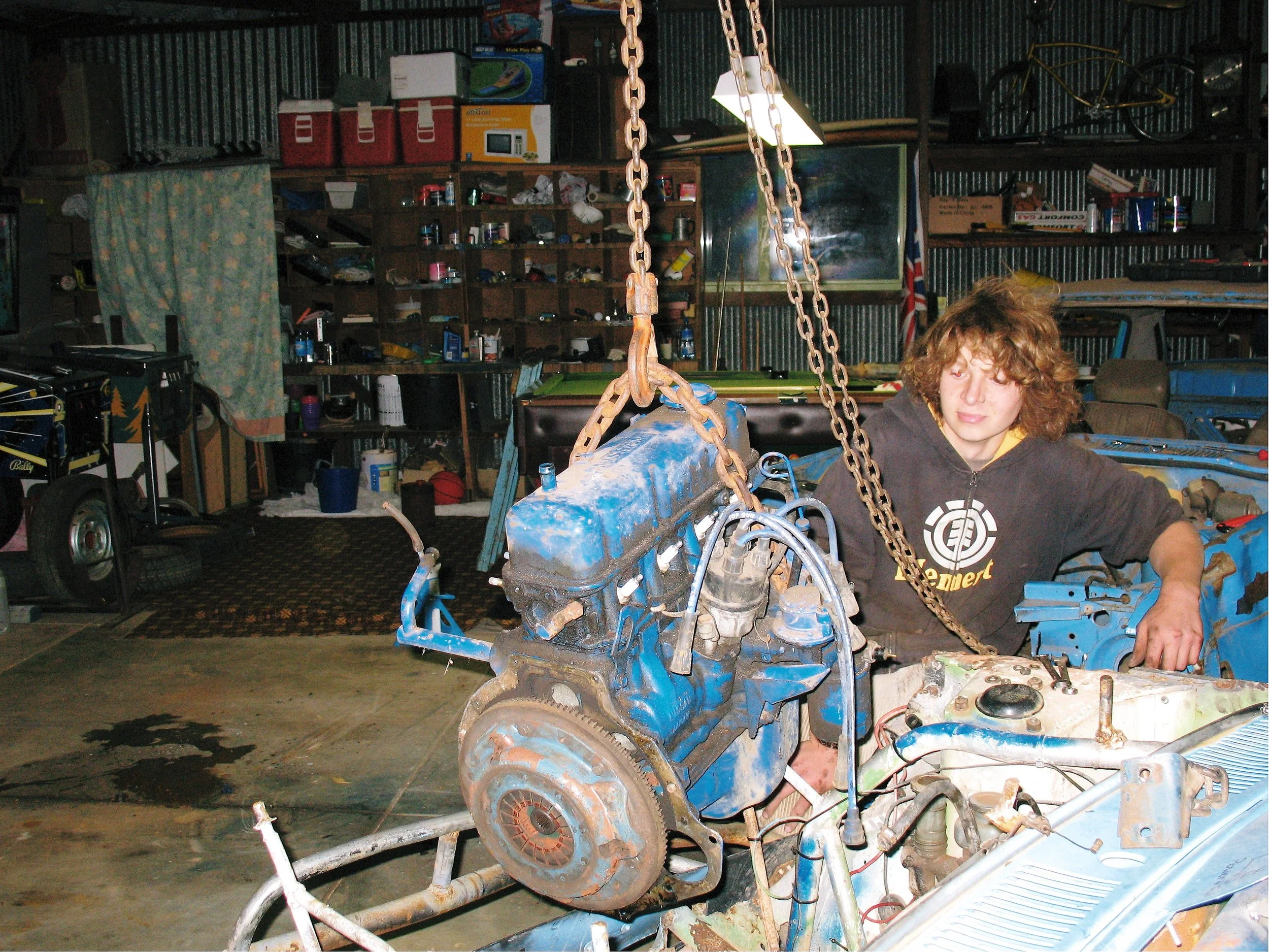 A person with curly hair working on a small engine in a cluttered garage or workshop. Chains are hanging from the ceiling, and shelves filled with various tools and supplies are visible in the background.