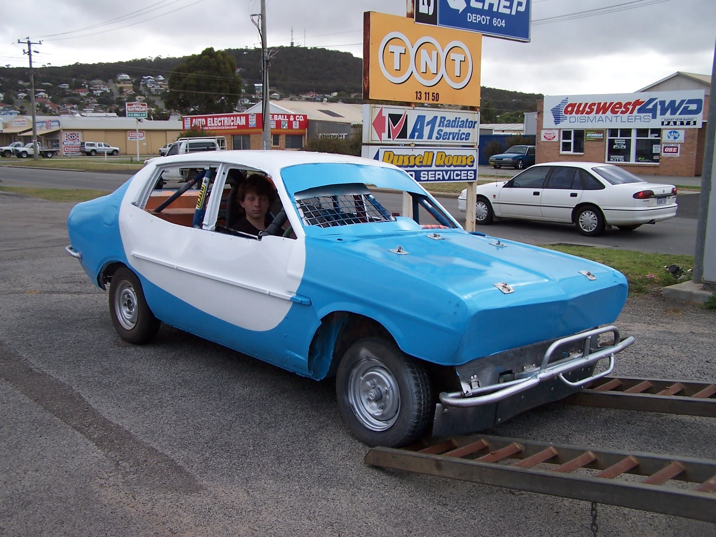 A young man sitting inside a blue and white race car with a roll cage on the street near a car parts and service shop, with signs for TNT, A1 Radiator Service, Russell Rouse, and AUSWEST Dismantlers visible in the background. The weather appears overcast.