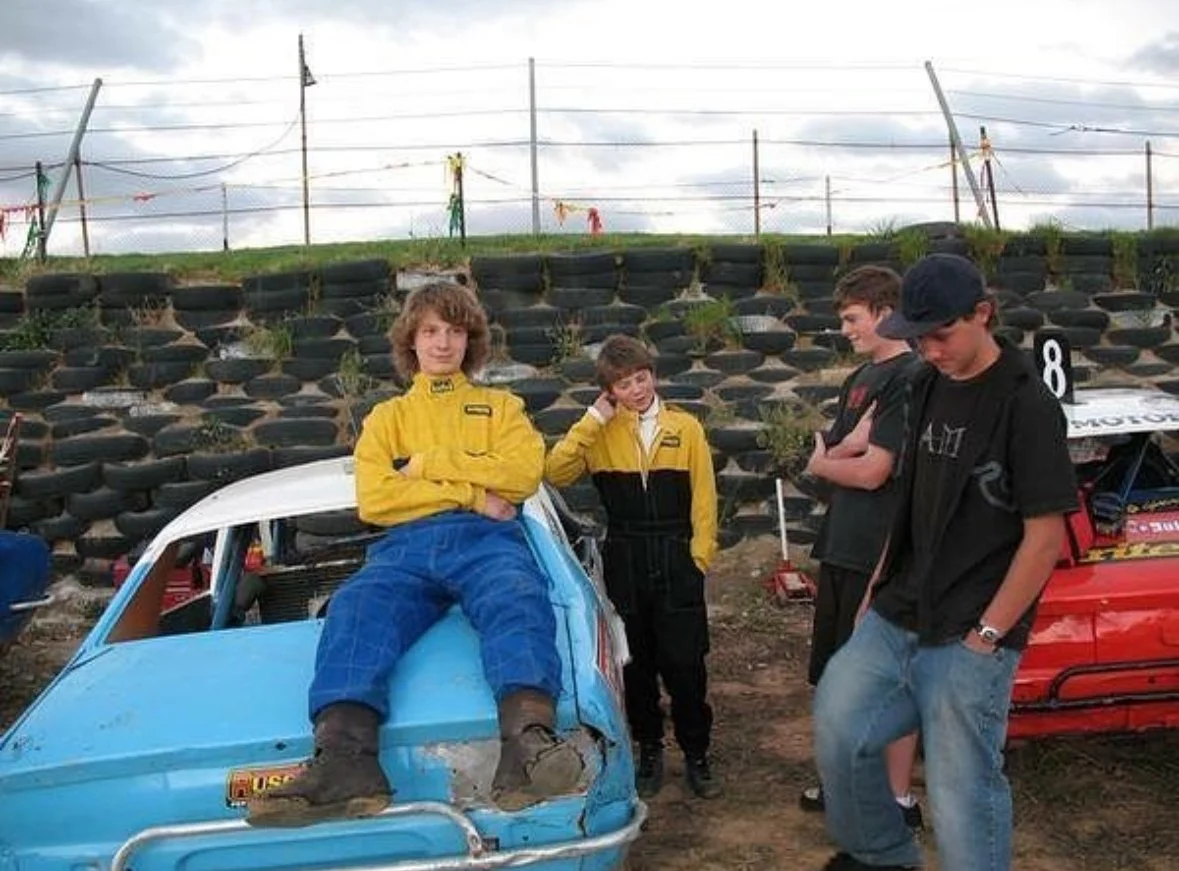 Four young boys at a racetrack with two race cars, one blue and one red. One boy in a yellow racing suit is sitting on the blue car, while the other boys stand nearby, some with arms crossed and one adjusting his headphones.