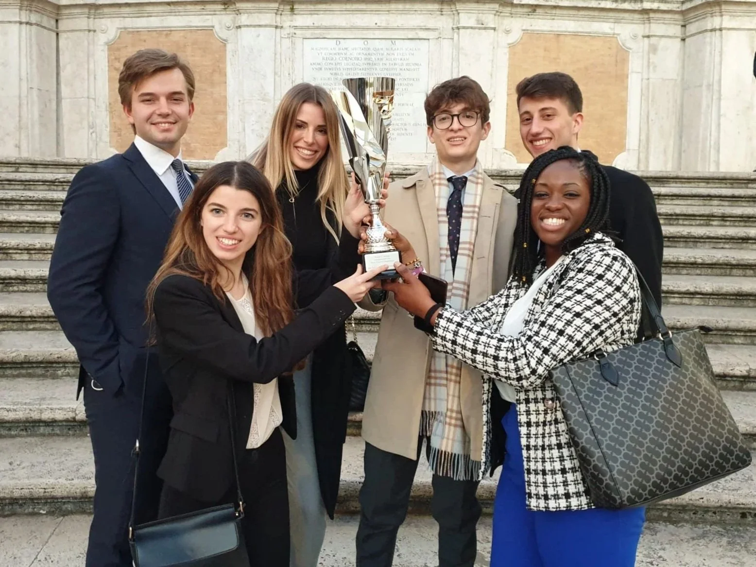 Group of six young adults standing on steps, holding a trophy together, smiling for the camera outdoors.