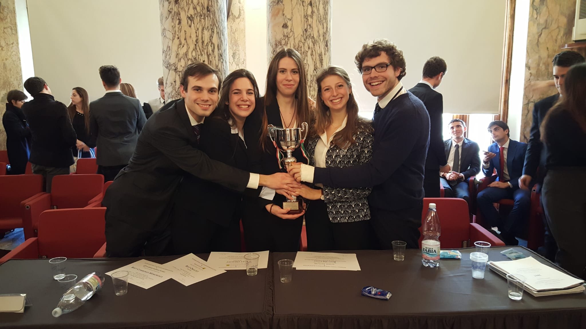 A group of five young adults in formal attire smiling and holding a trophy, standing in a room with patterned marble columns and large windows with roller shades, while other people are standing and sitting in the background.