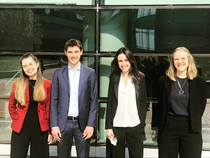 Four young adults standing outdoors in front of a modern glass building, smiling for the photo.