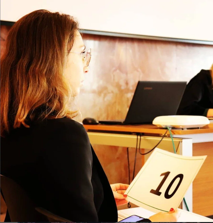 A woman with reddish-brown hair, glasses, and a black blazer sitting at a table in a classroom or office setting, holding a white sign with the number 10 on it, with a laptop and other office equipment on the table.