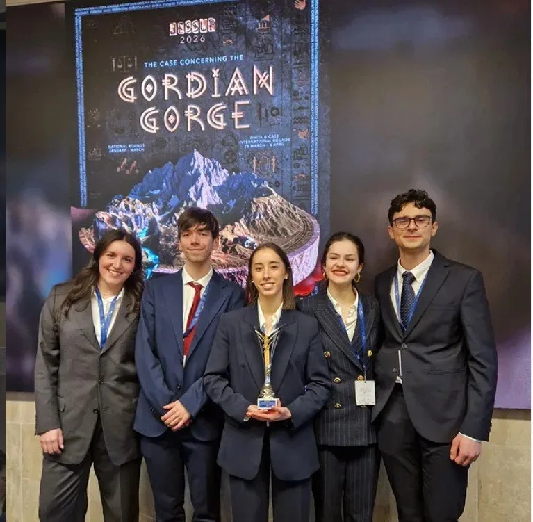 Group of five young people in formal attire standing and smiling in front of a poster titled 'The Case Concerning the Gordian Gorge'. The woman in the center is holding a trophy.