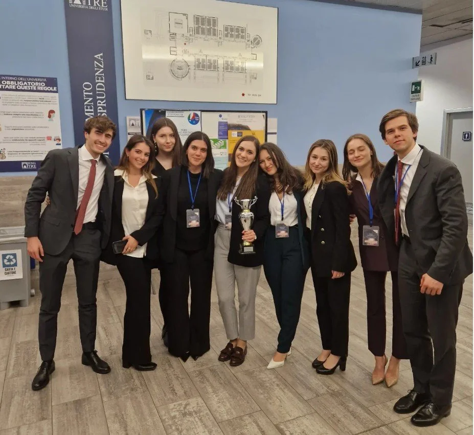 A group of nine young adults in business attire, standing together indoors, holding a trophy, and smiling.