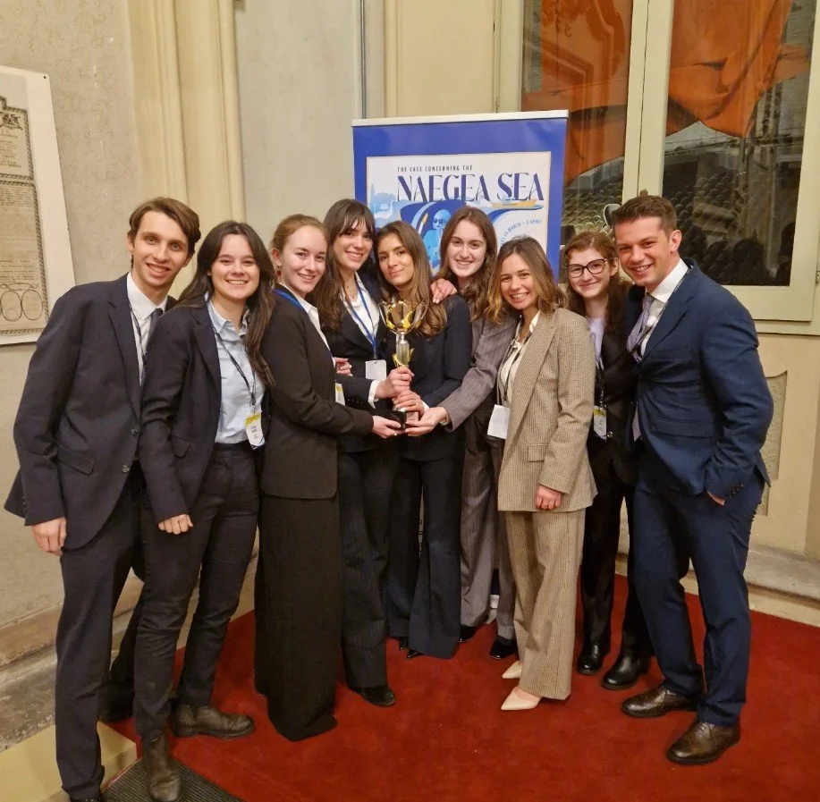 Group of young adults in formal attire holding a trophy at an event celebrating the voyage to the NAEGEA Sea, with a blue banner and historic building interior in the background.