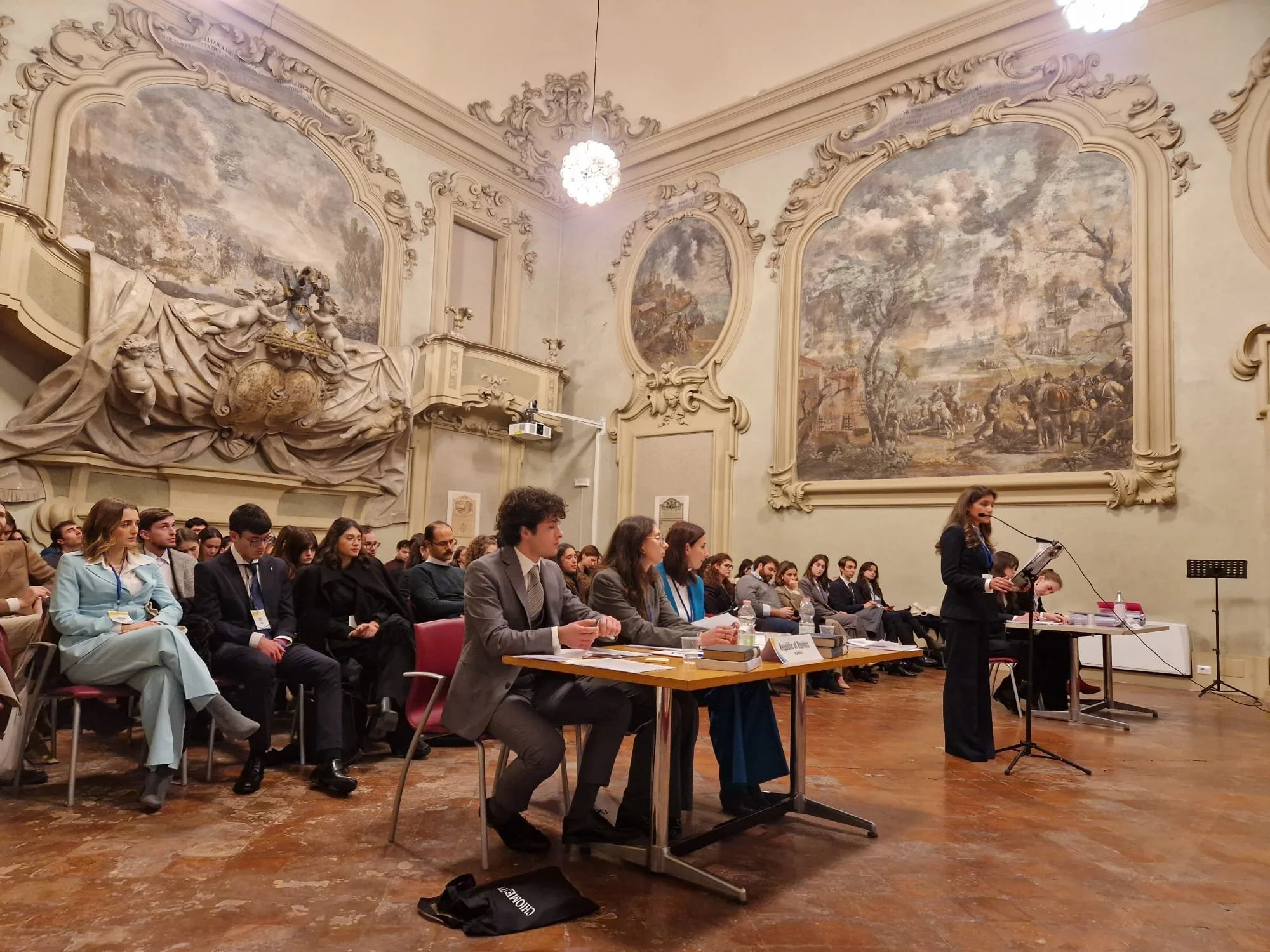 A large gathering of young adults seated in a formal room with ornate wall murals and decorative sculpture. A woman is speaking at a podium with a microphone, and some individuals at a table in front are taking notes.