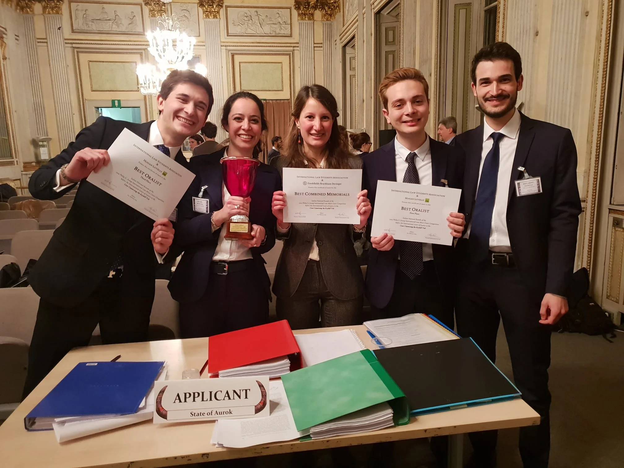 Group of five young adults dressed in formal attire, smiling and holding certificates and a trophy at an awards ceremony inside a lavish, gold-accented hall with chandeliers. In the foreground, there is an applicant registration table with folders and a sign labeled 'Applicant'.