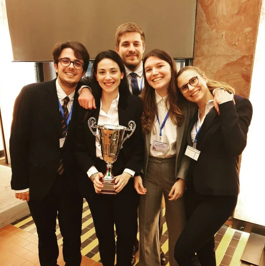 Group of five young adults in formal business attire celebrating at an event, with one woman holding a large trophy.