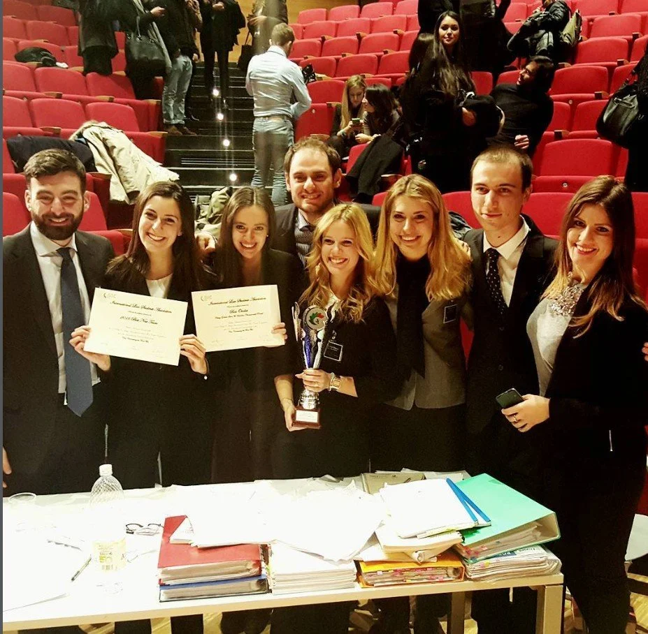 A group of eight young people in formal attire posing with certificates and a trophy, in an auditorium with red seats.