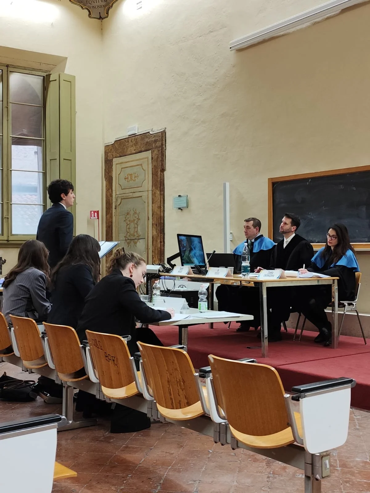 A courtroom scene with a young man standing and speaking to three judges sitting at a desk, some protesters or observers sitting in chairs, and a blackboard in the background.