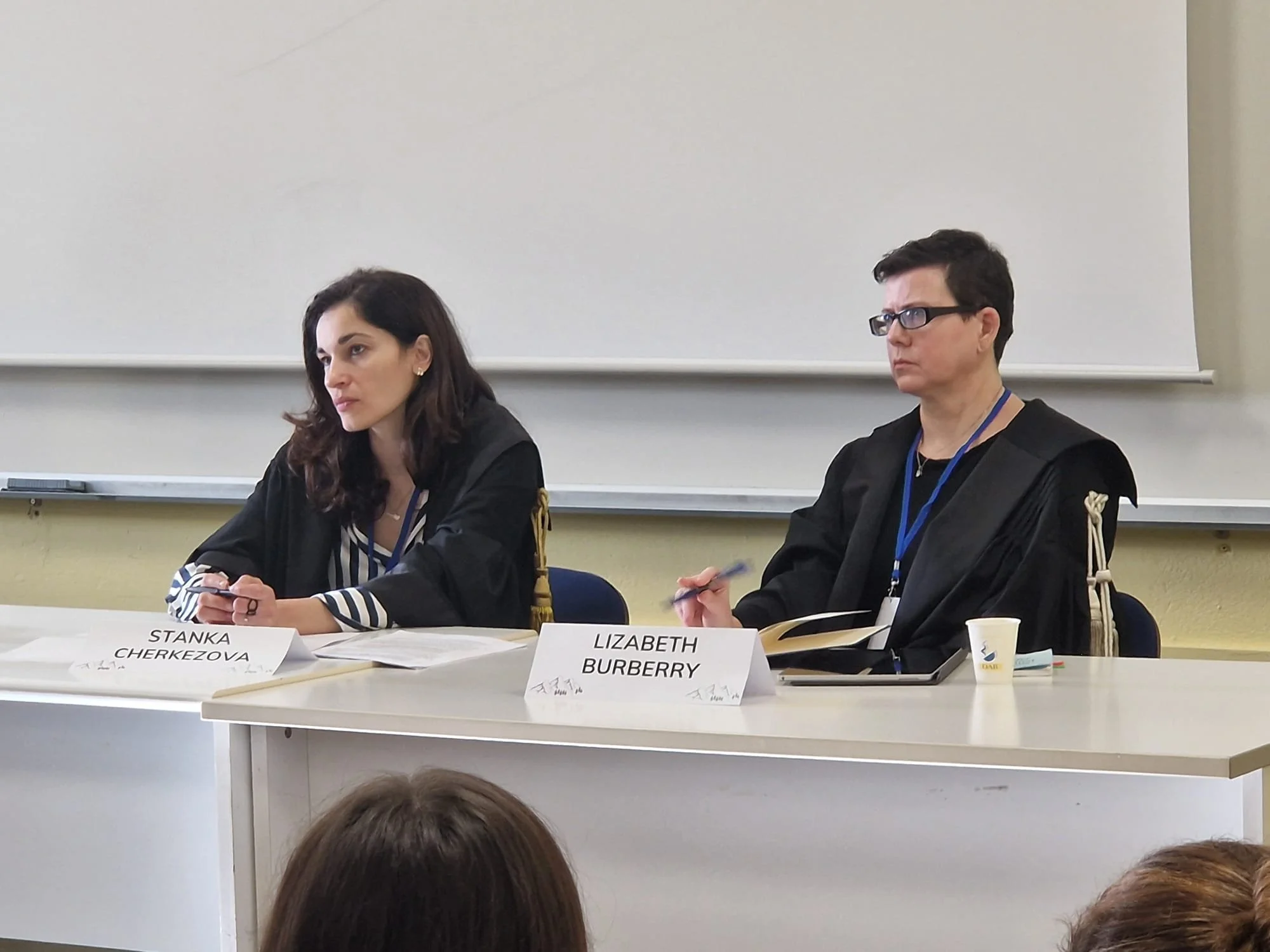 Two women sitting at a table during a formal event or conference, with name tags reading 'Stanka Cherkezova' and 'Elizabeth Burberry' in front of them, against a plain wall and whiteboard in the background.